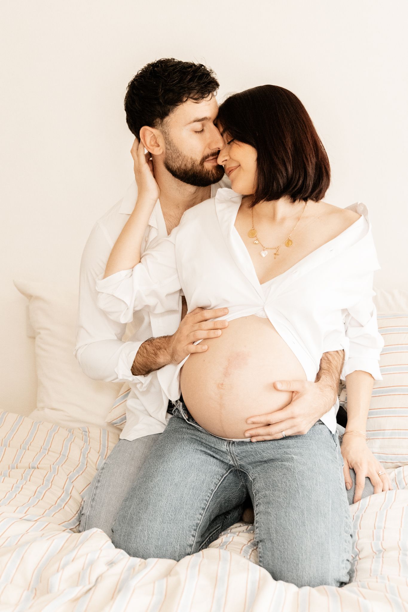 Couple sharing a tender kiss on bed, man embracing pregnant woman showing bare baby bump.
