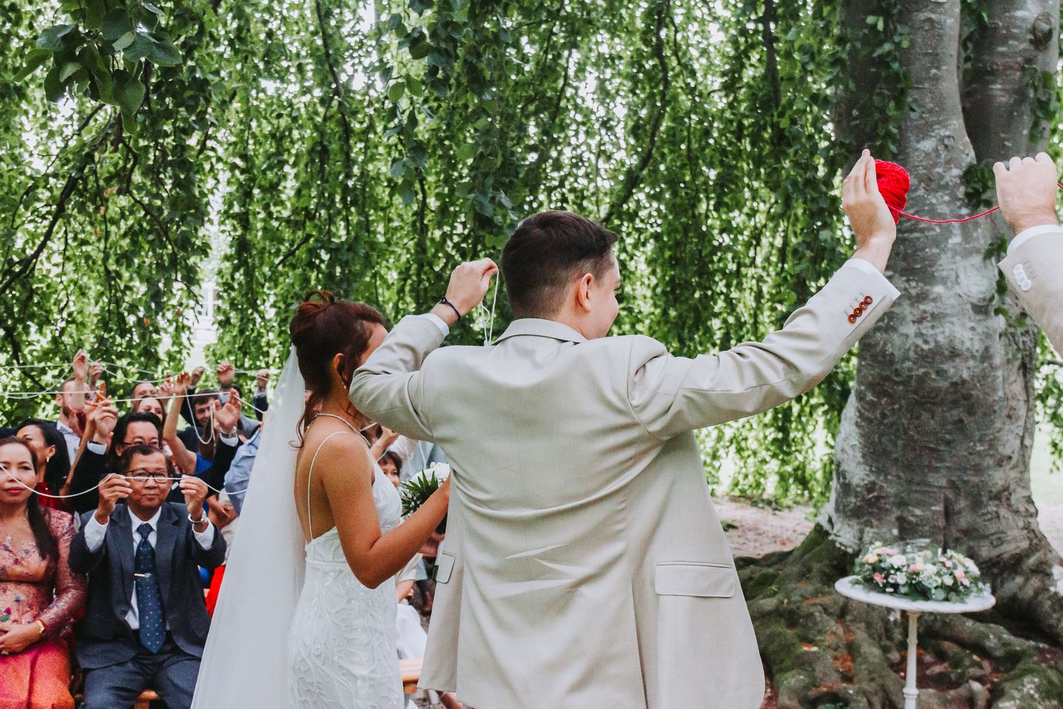 Newlywed couple celebrating with raised hands facing cheering guests under weeping willow wedding ceremony.