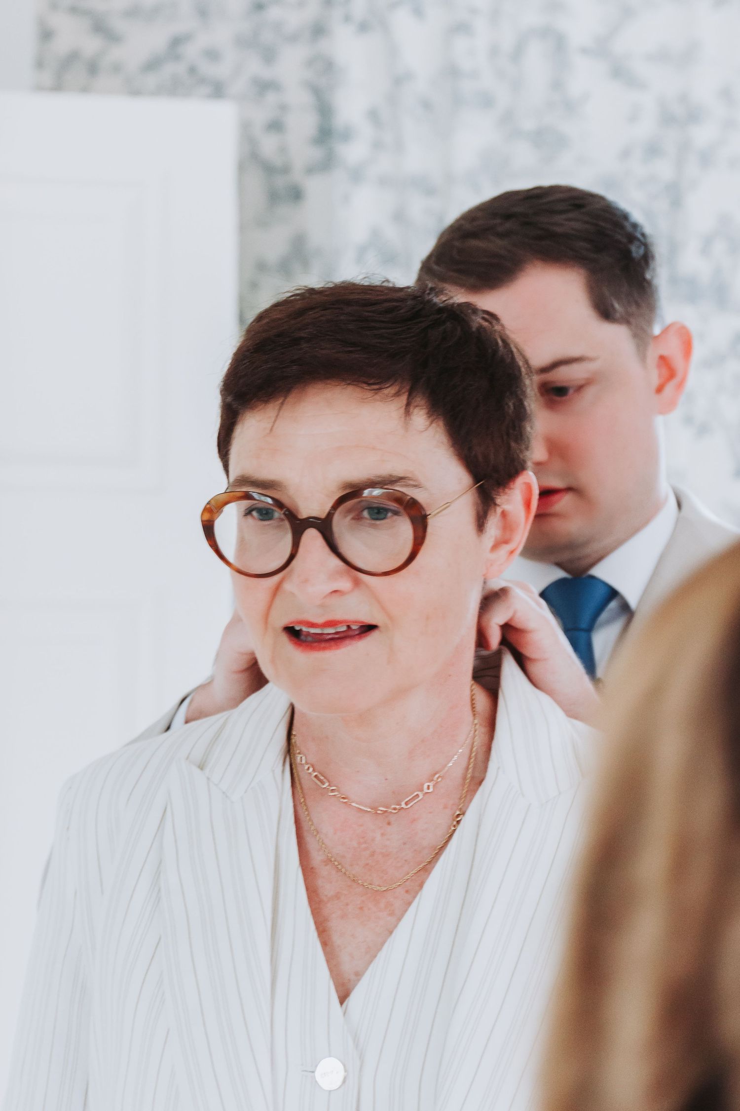 Young man adjusting a necklace on an older woman with short hair and round tortoiseshell glasses.