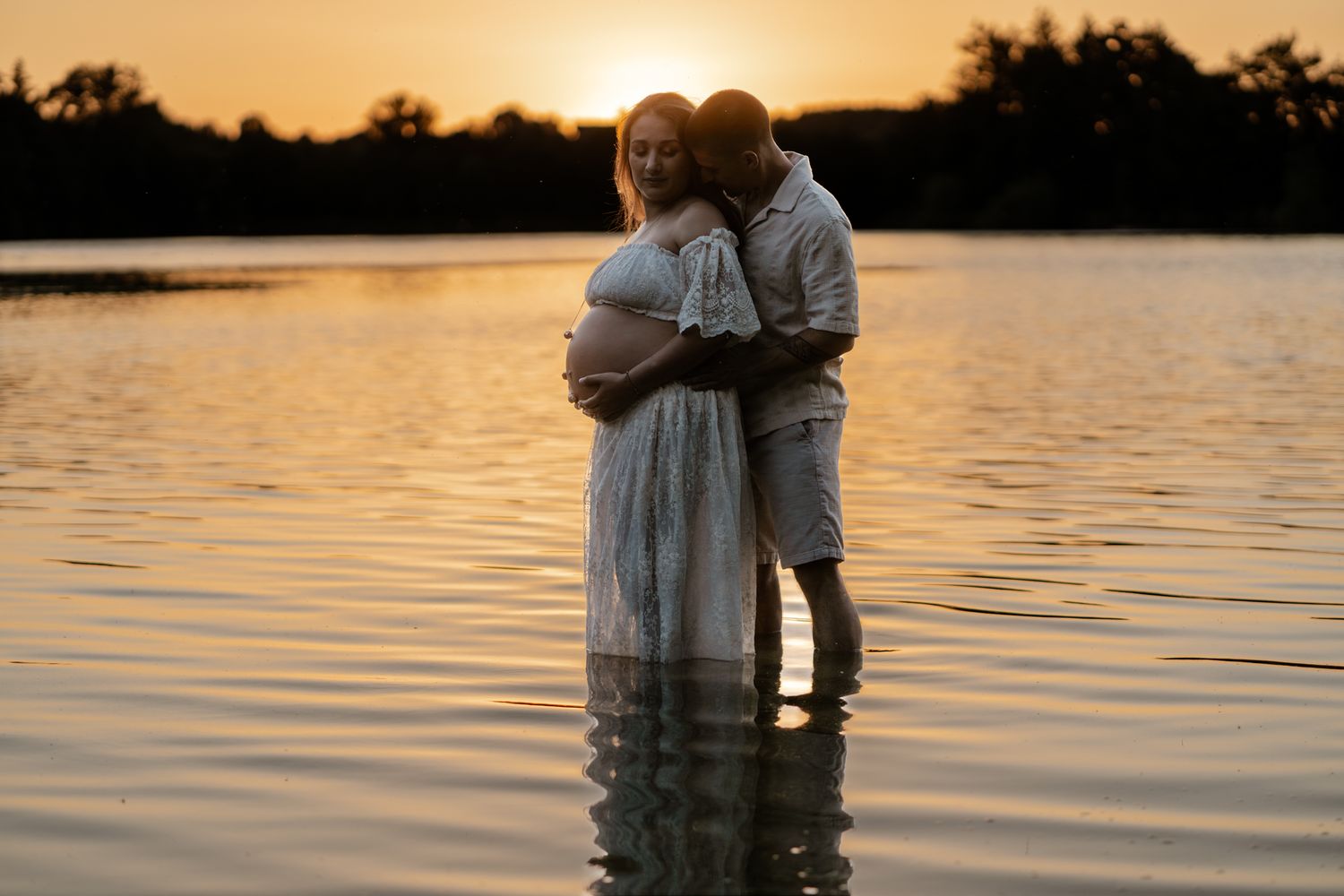 Pregnant couple embracing in a lake at golden sunset, woman in white dress cradling baby bump.