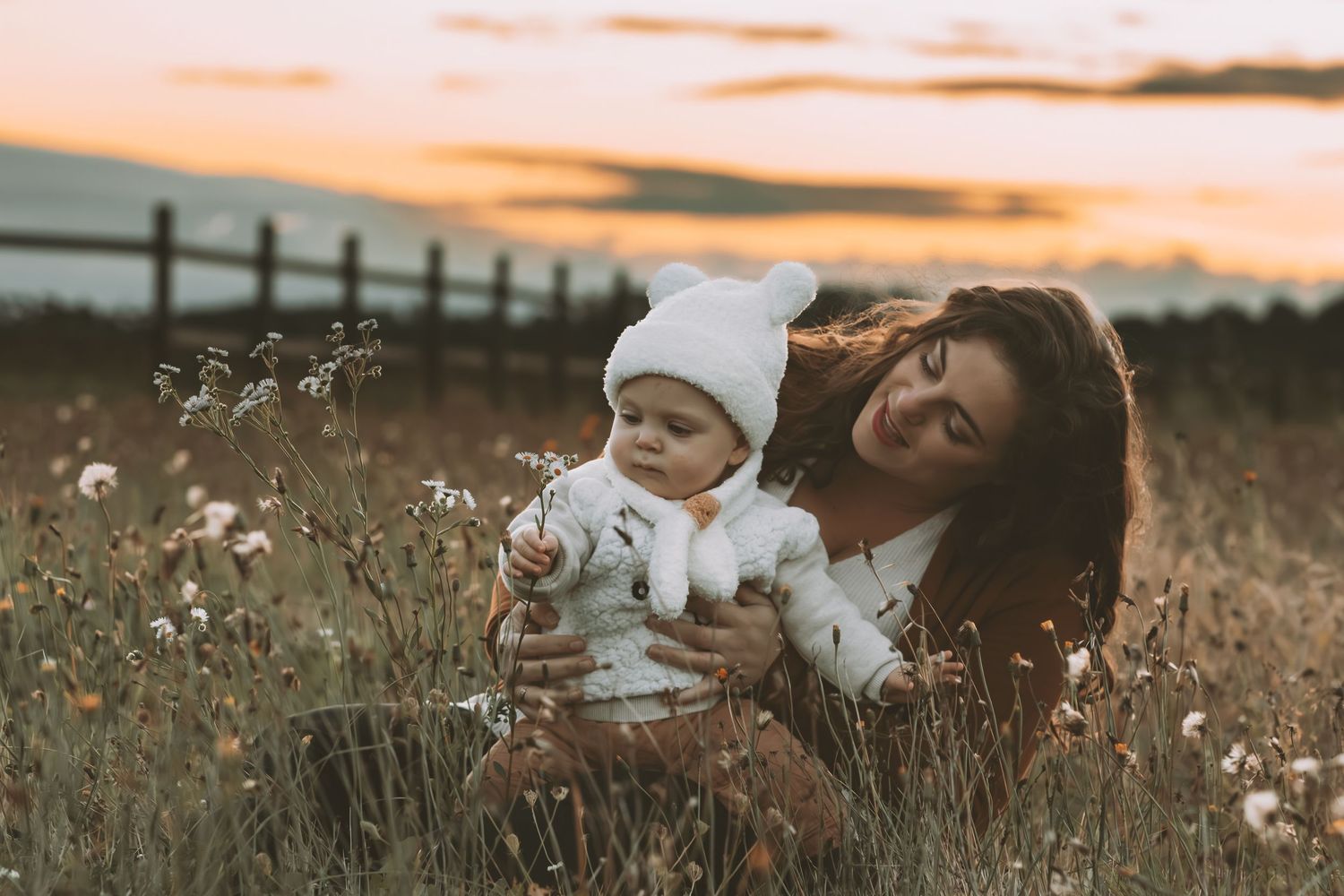 Mother and baby sitting in wildflower field at sunset, sharing a peaceful moment together.