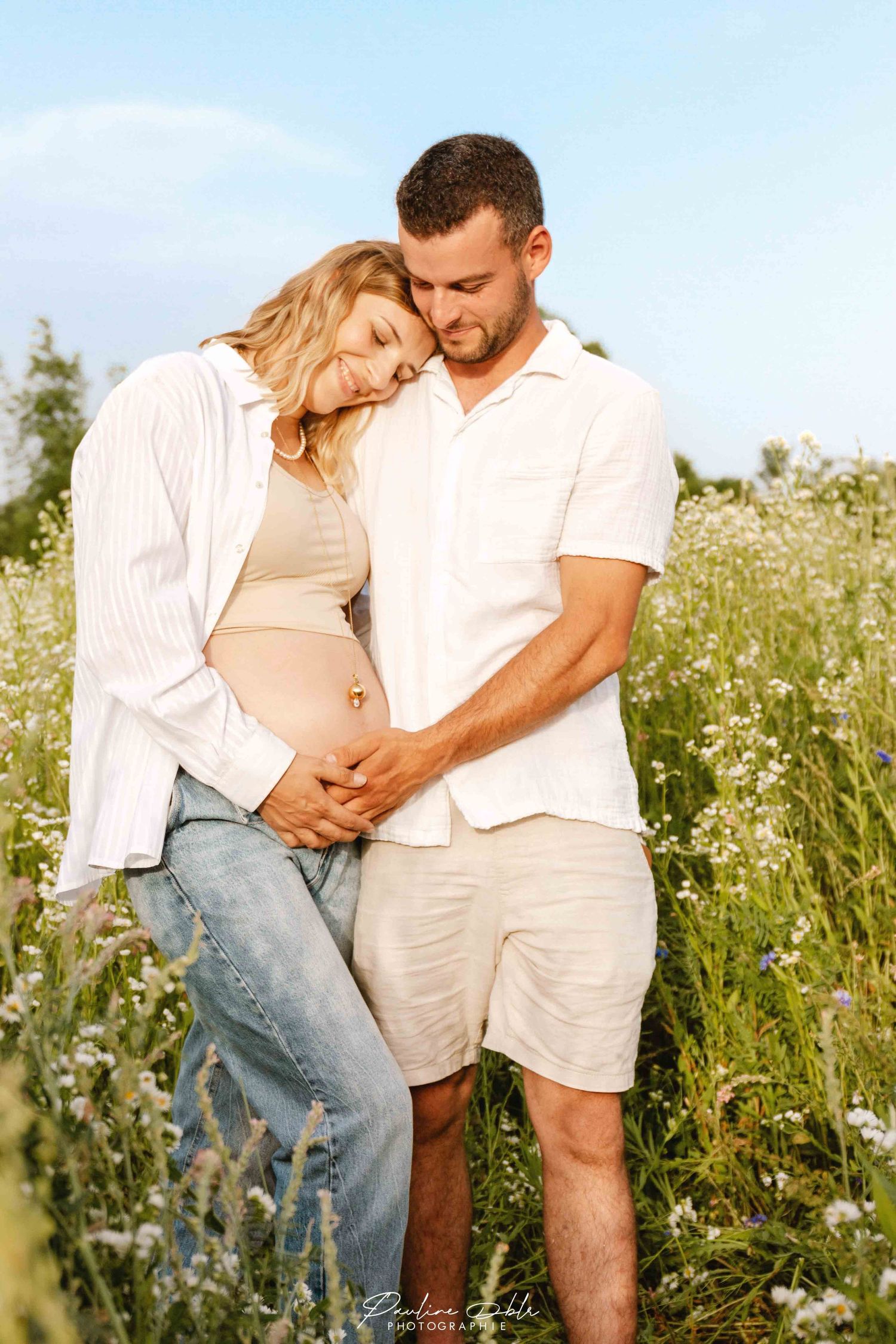 Couple embracing in a wildflower field at golden hour, hands cradling pregnant belly.