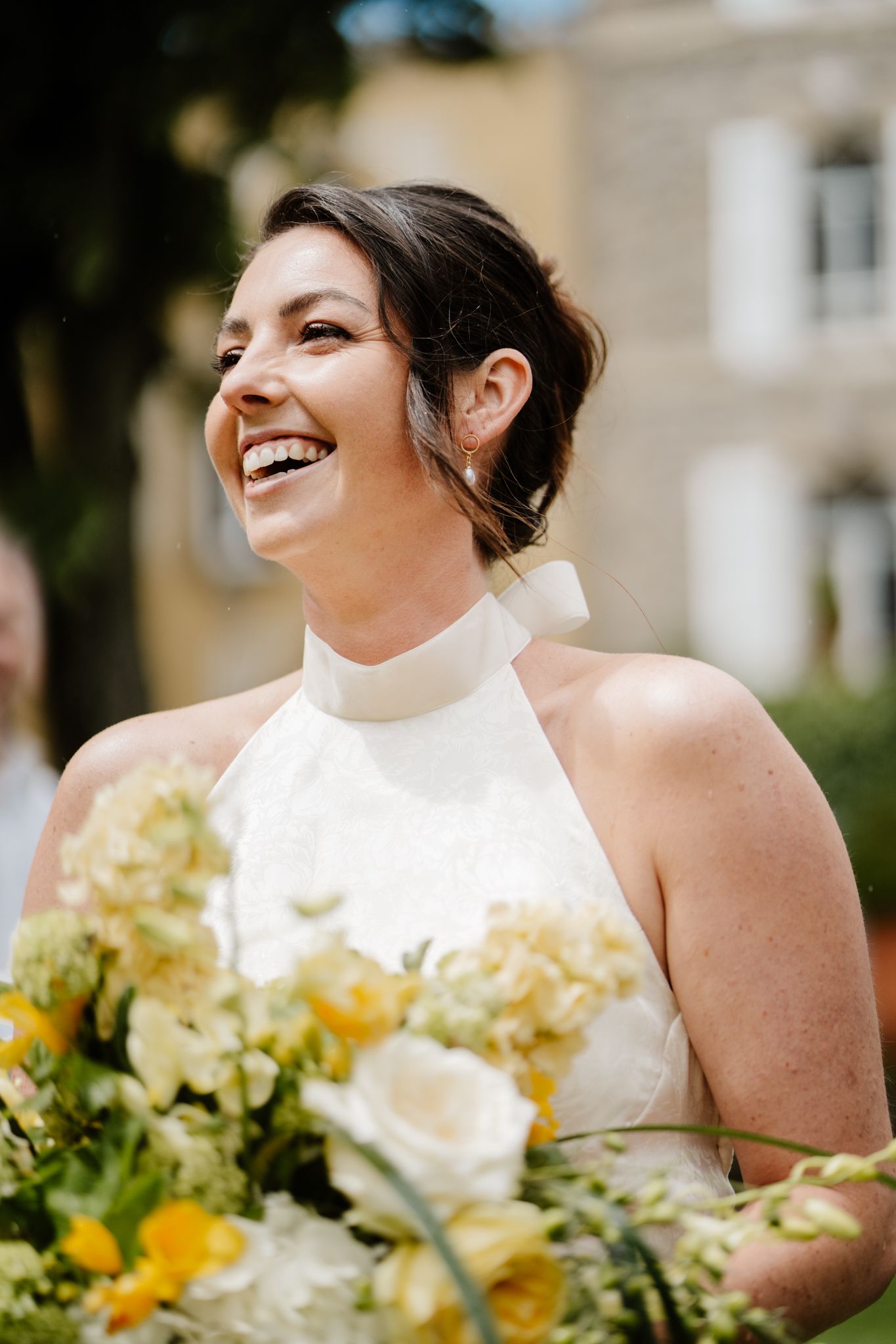 Radiant bride in white halter gown laughing joyfully while holding yellow and white floral bouquet.