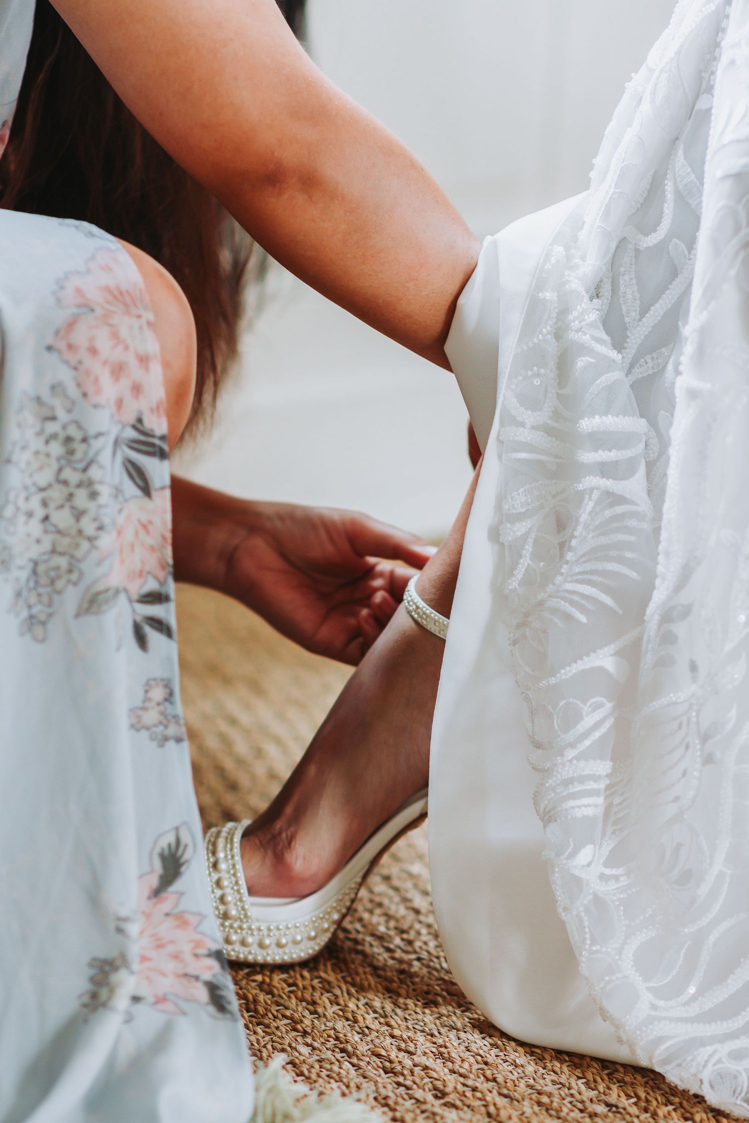 Bridesmaid fastening pearl-adorned white bridal heels on the bride's ankle beside a lace wedding gown.