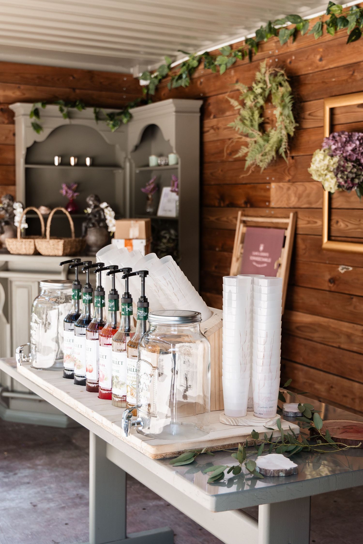Elegant syrup bar station with flavored bottles and dispensers on a table in a rustic wood-paneled wedding venue.