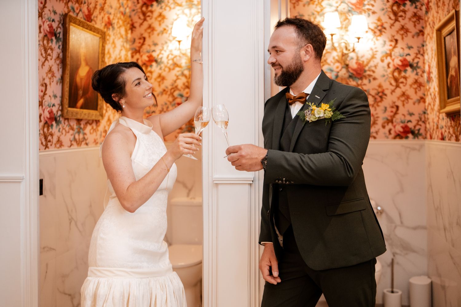 Bride and groom toasting champagne in a floral wallpapered room, groom wearing green suit with bow tie.