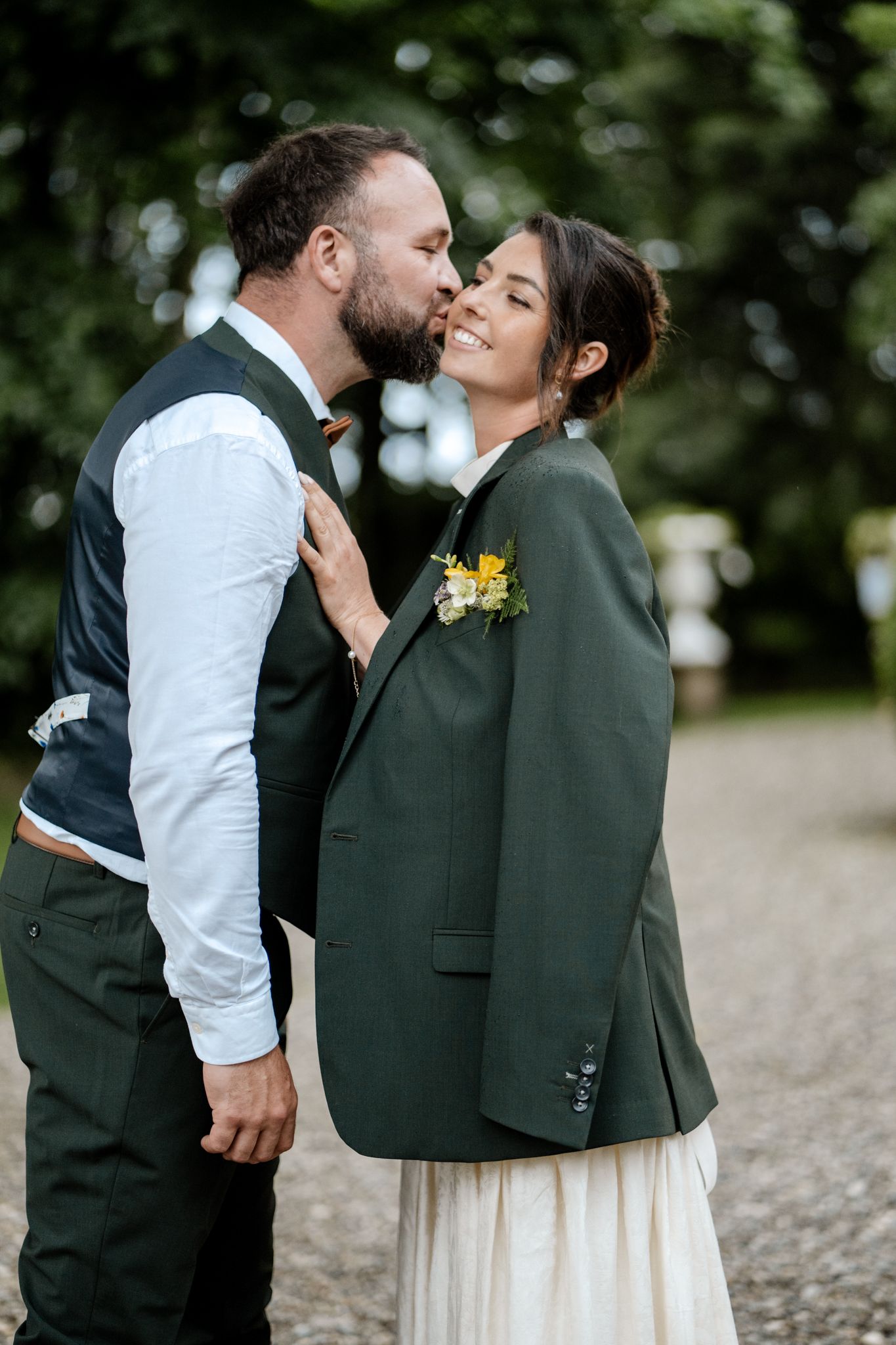 Groom kissing smiling bride on cheek outdoors, bride wearing groom's green jacket over wedding dress.