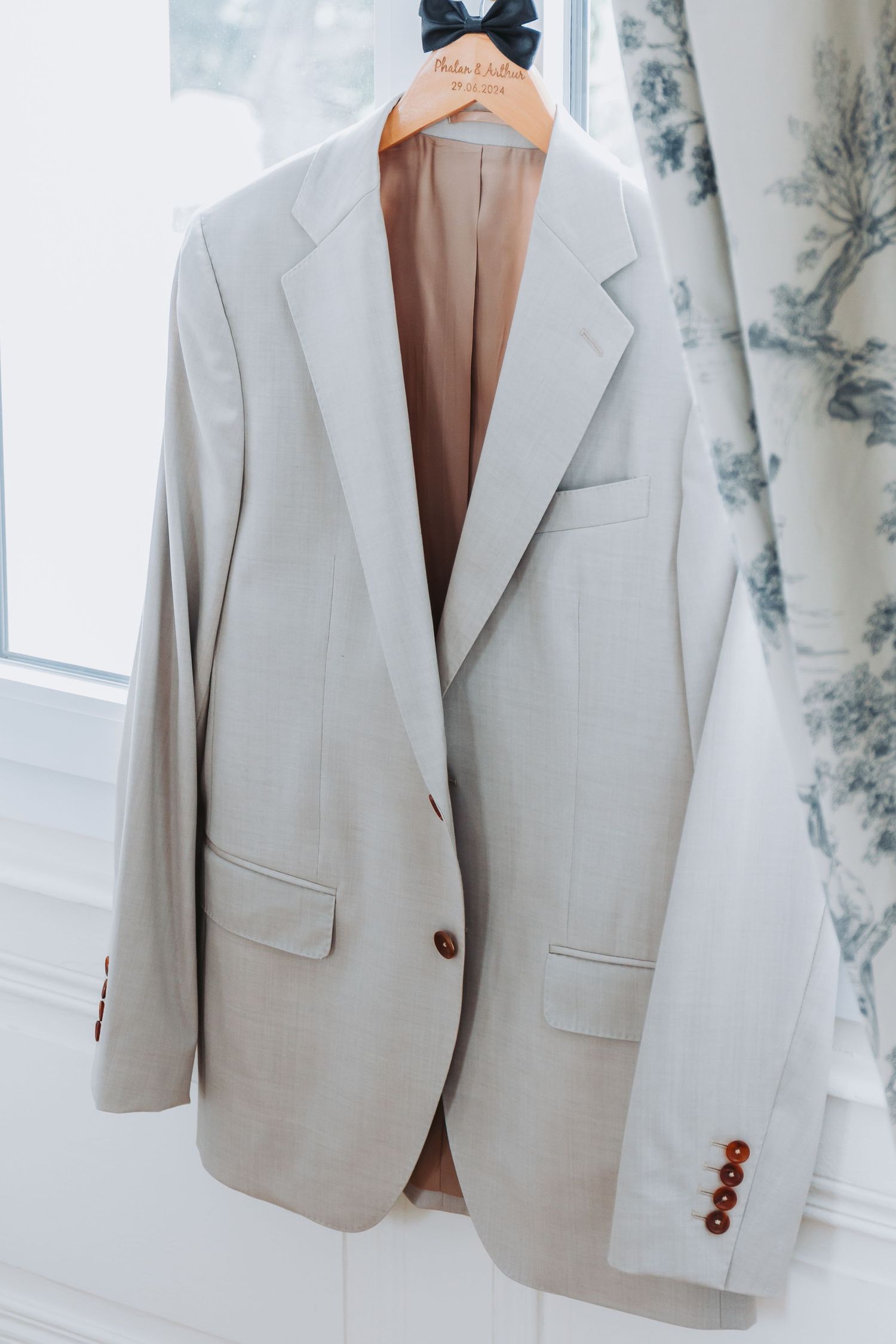 Light beige groom suit jacket hanging on a personalized wooden hanger by a bright window.