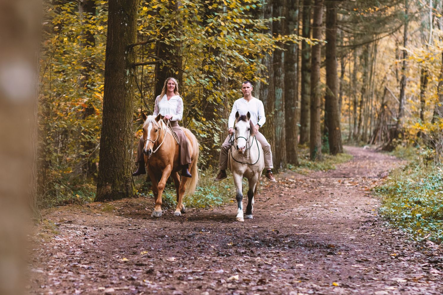 Two riders in white shirts on horseback trotting through a colorful autumn forest trail.