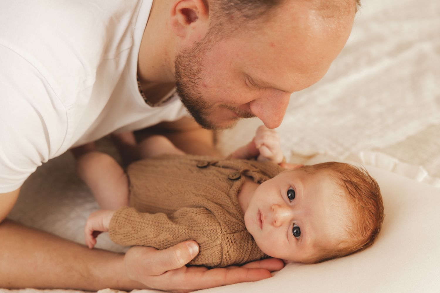 Loving father in white t-shirt leaning down to kiss his newborn baby lying on a white blanket.