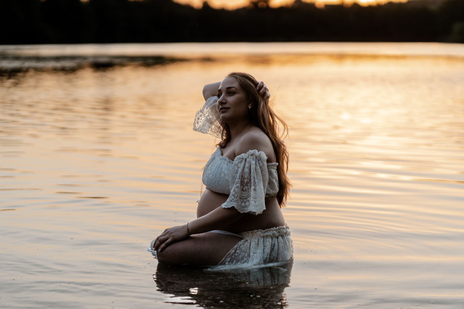 Pregnant woman in white lace outfit sitting gracefully in lake water during a glowing sunset.