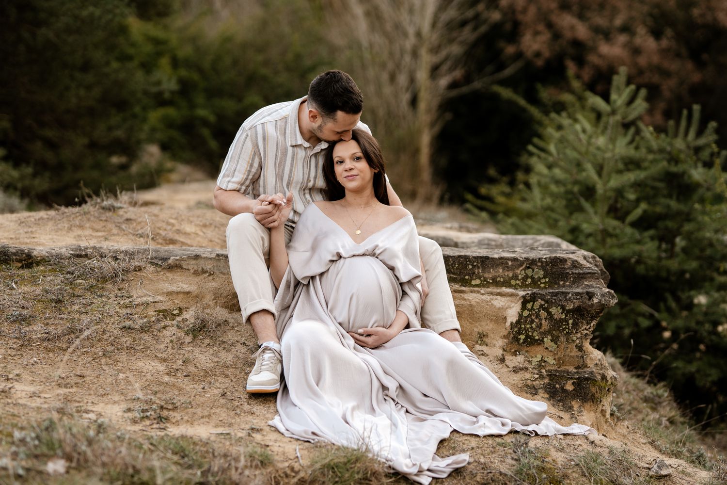Séance photo grossesse en Isère : un futur papa embrasse la tête de la future maman en robe nacrée qui berce son ventre.