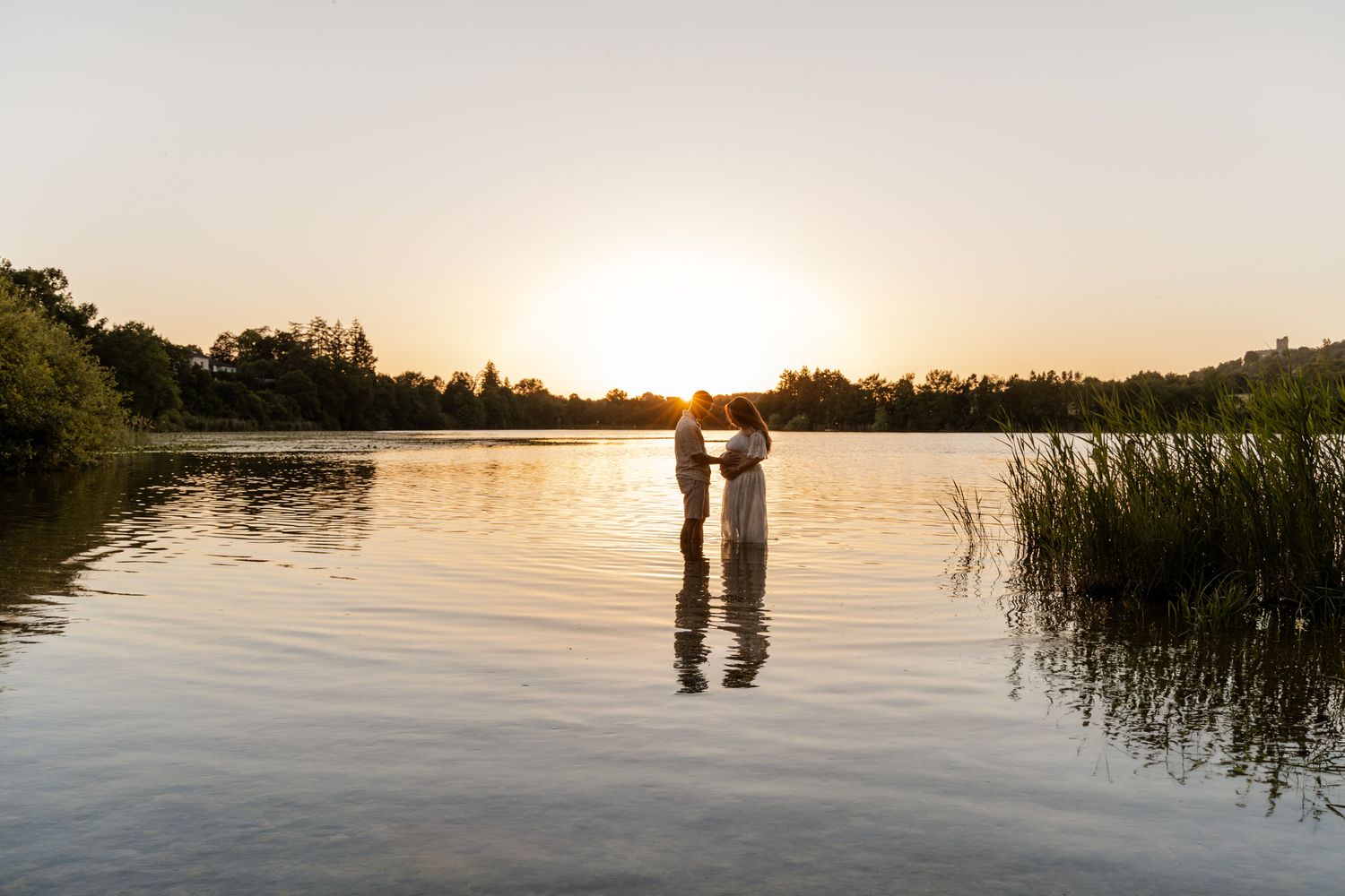 Couple holding hands standing in calm lake water with vibrant golden sunset and lush greenery behind.