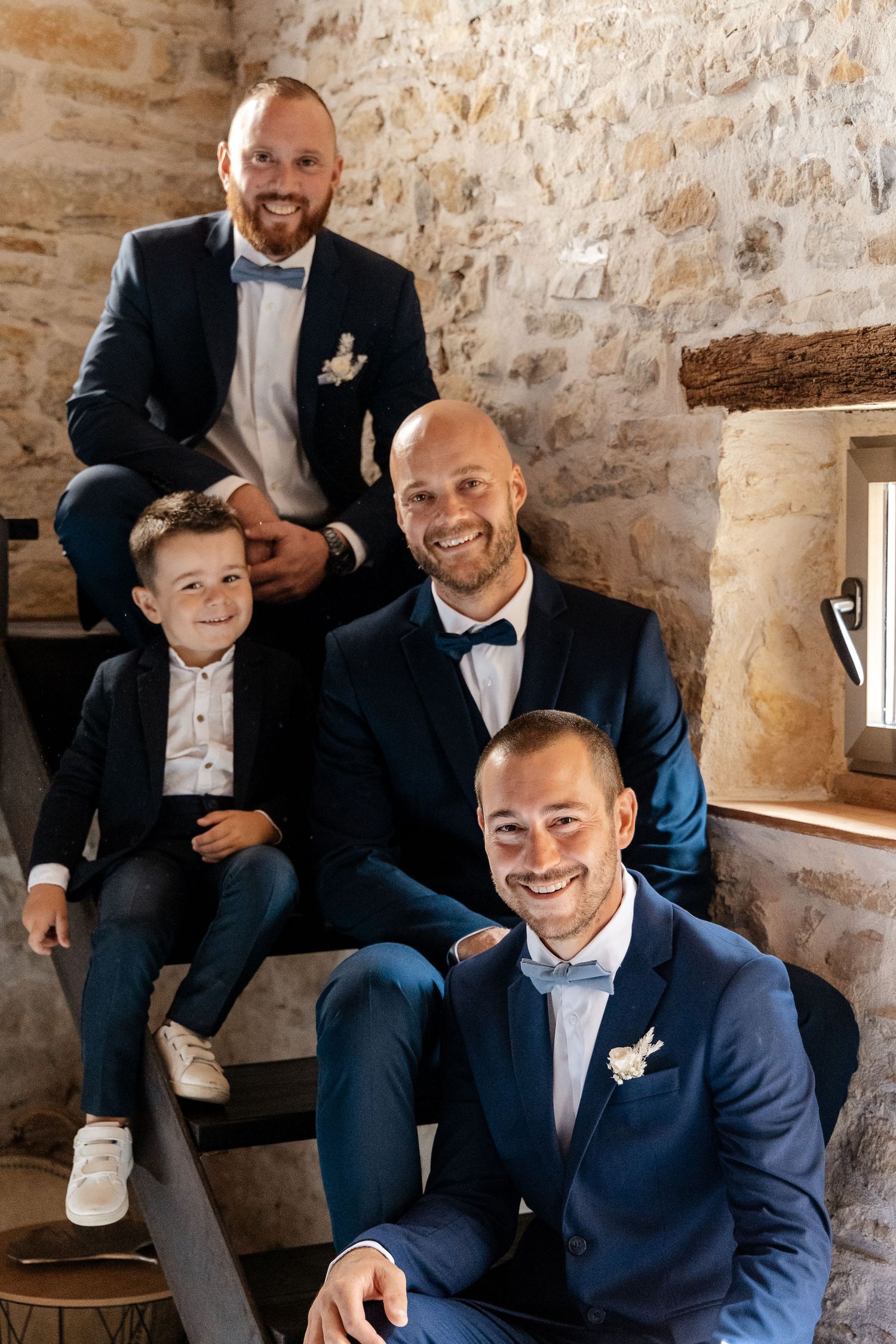 Four smiling males in navy wedding suits posing on rustic stone staircase indoors.