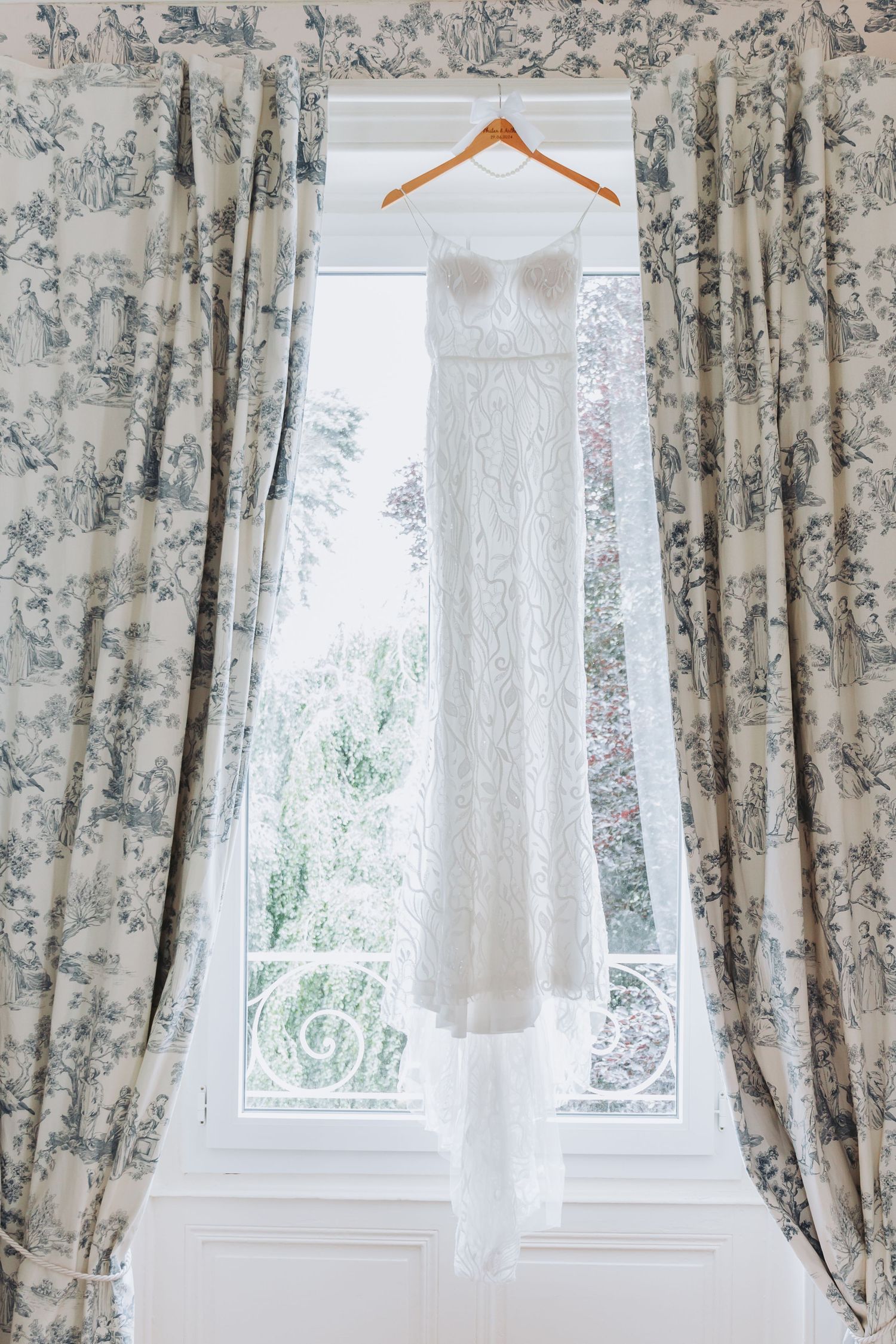 Elegant white lace wedding dress hanging on a wooden hanger in front of a bright window with floral curtains.