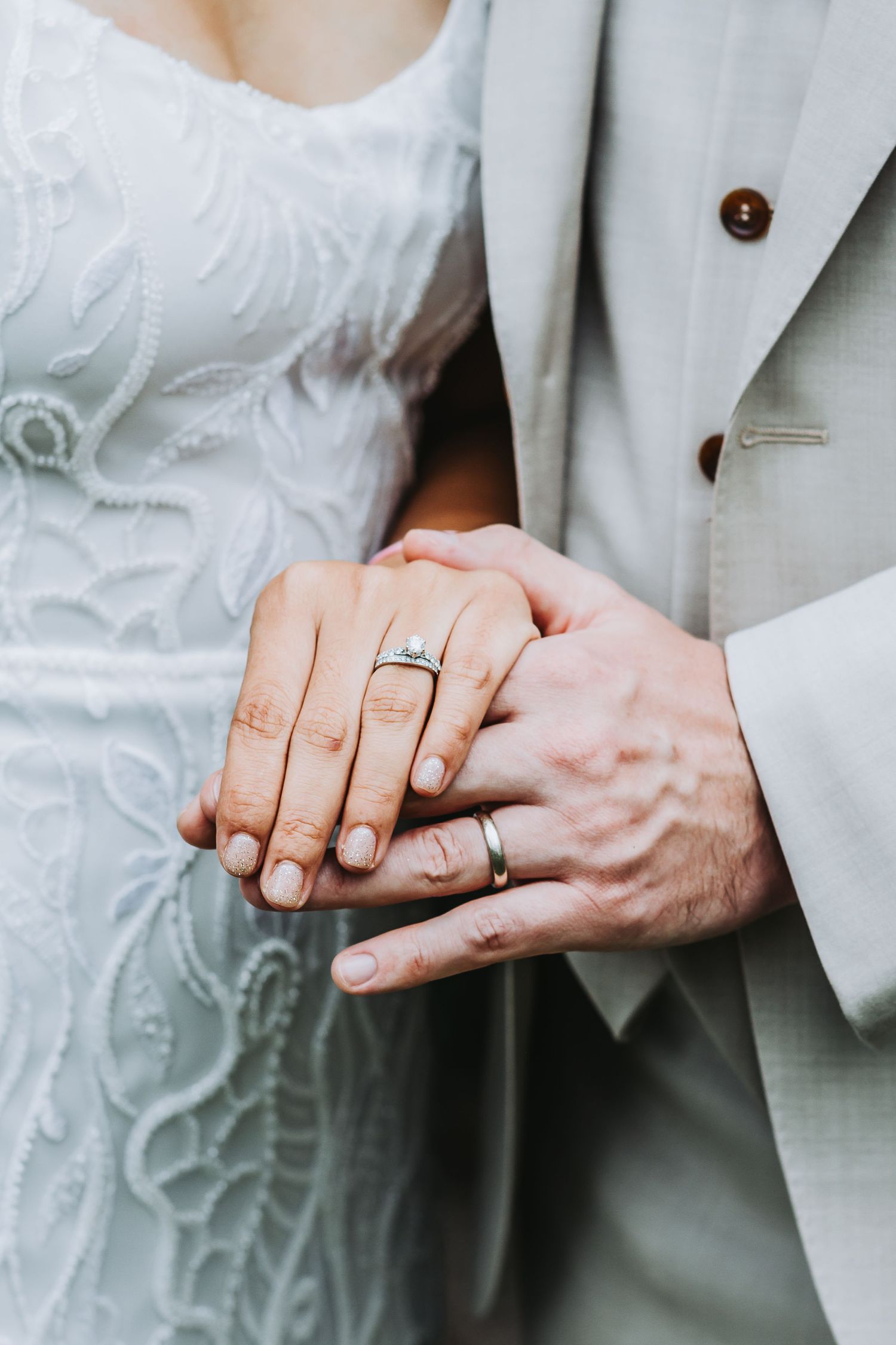 Close-up of newlyweds holding hands showing wedding rings, bride in white lace dress and groom in beige suit.