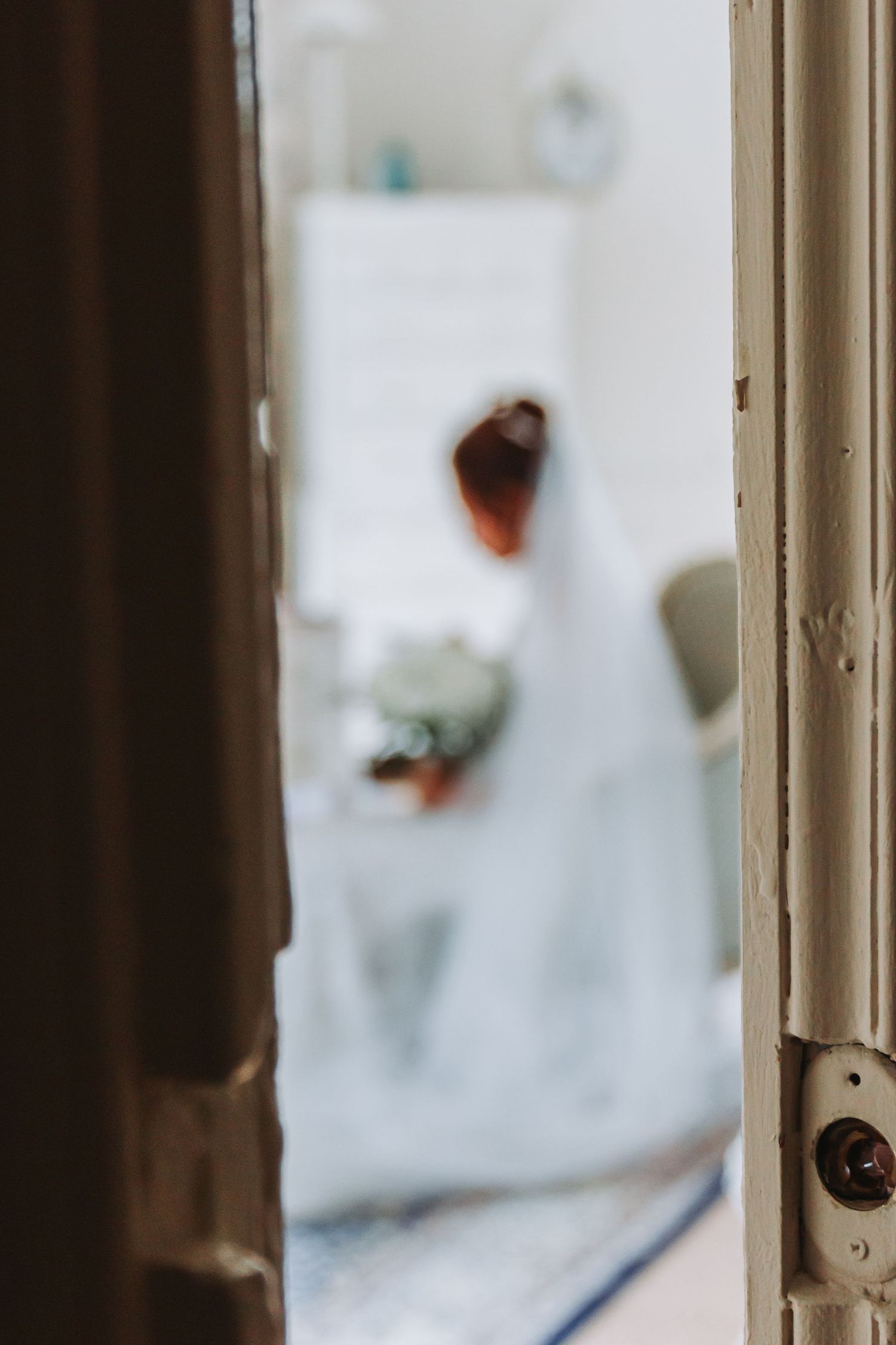 Blurred bride in white wedding dress and veil holding bouquet viewed through vintage doorway.