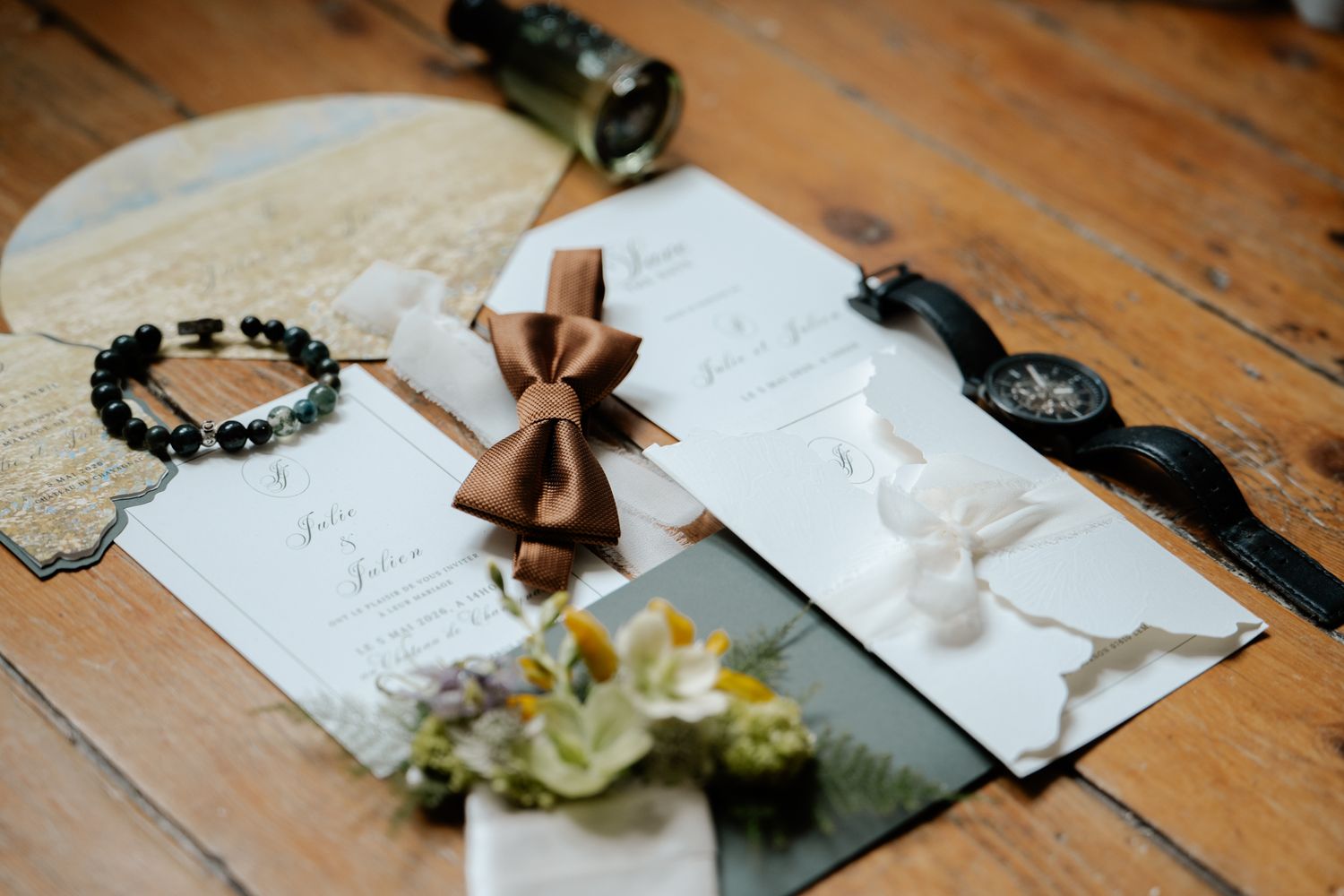 Wedding stationery flat lay featuring brown bow tie, invitation cards, watch, bracelet, and yellow flowers.