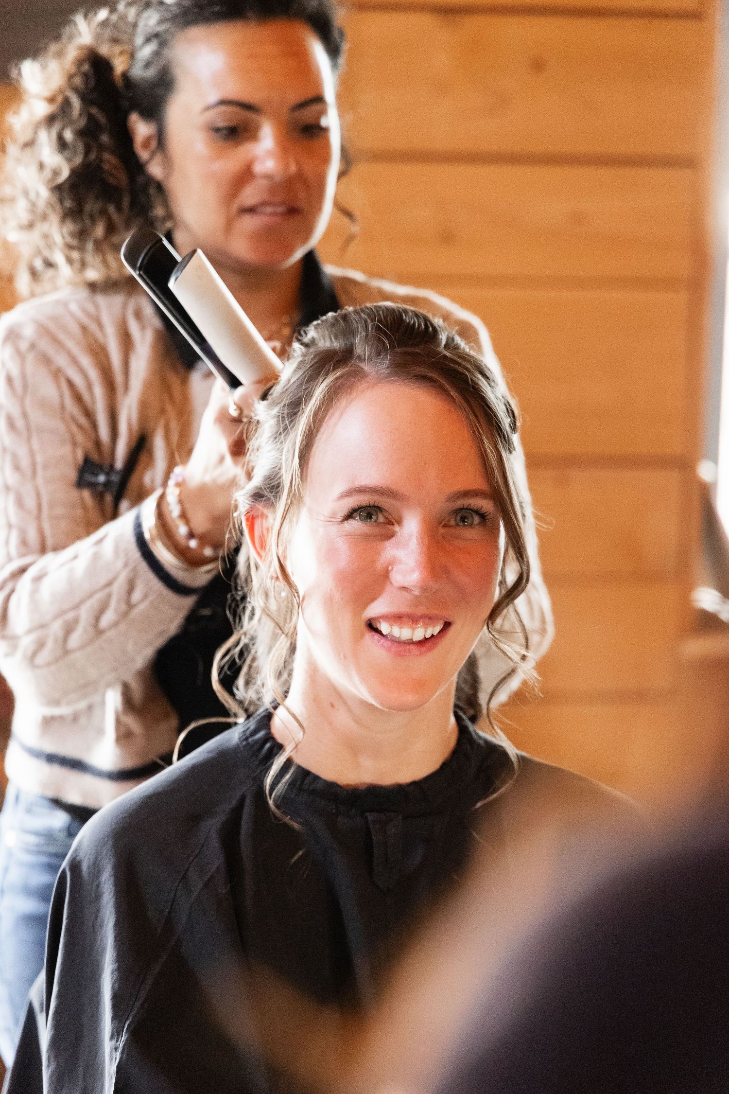 Smiling bride getting her hair styled with a curling iron by a hairdresser in a wooden cabin.