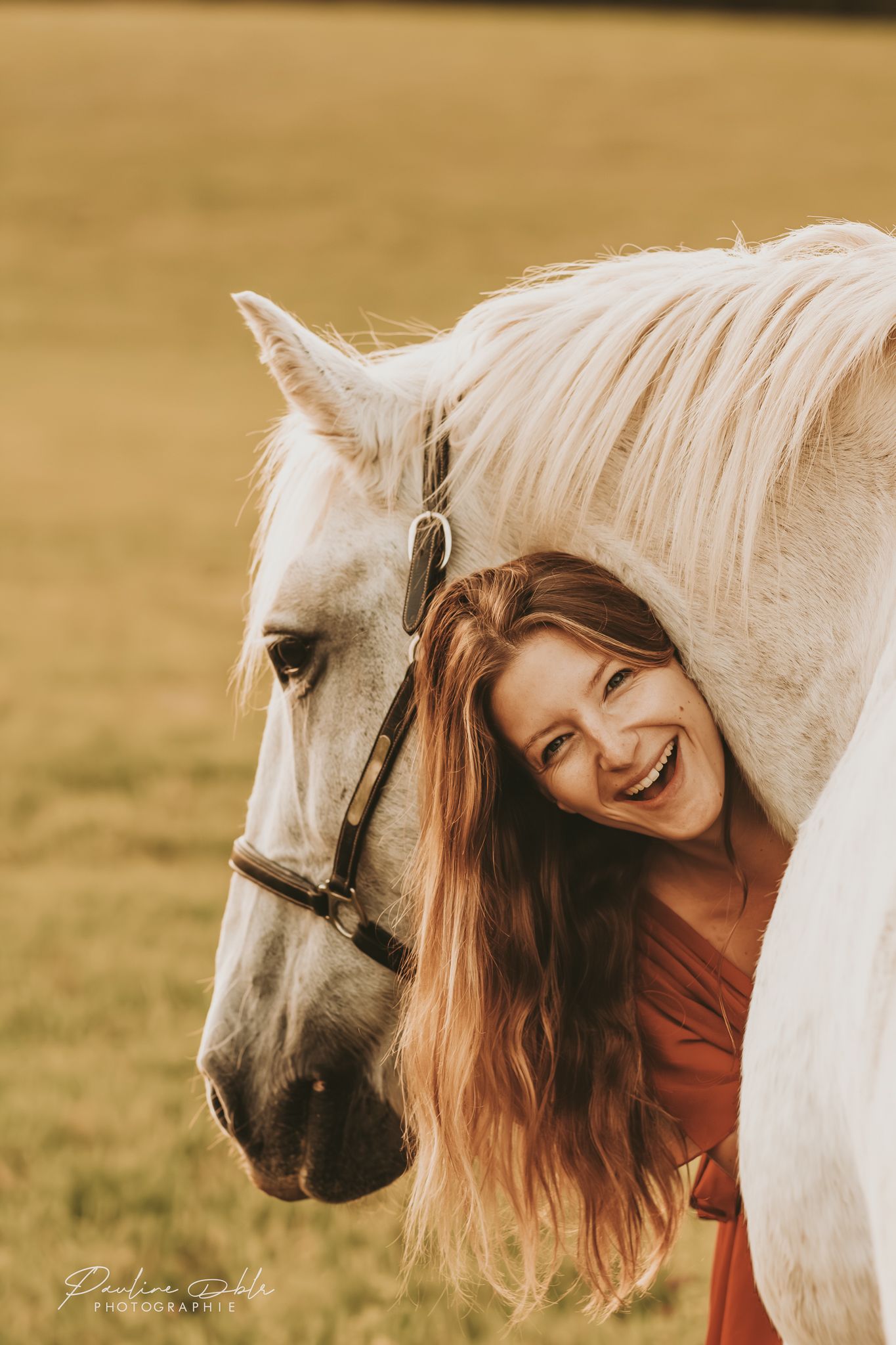 A laughing woman with red hair hugs a white horse in a golden field.