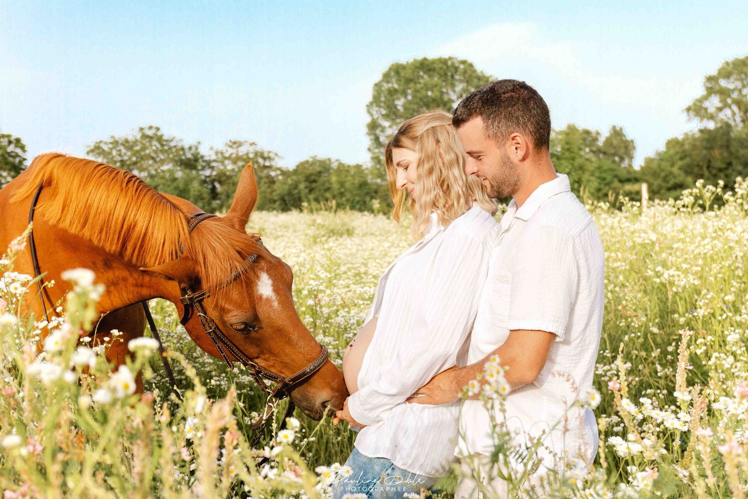 Couple in white standing in a wildflower field with a chestnut horse on a sunny day.