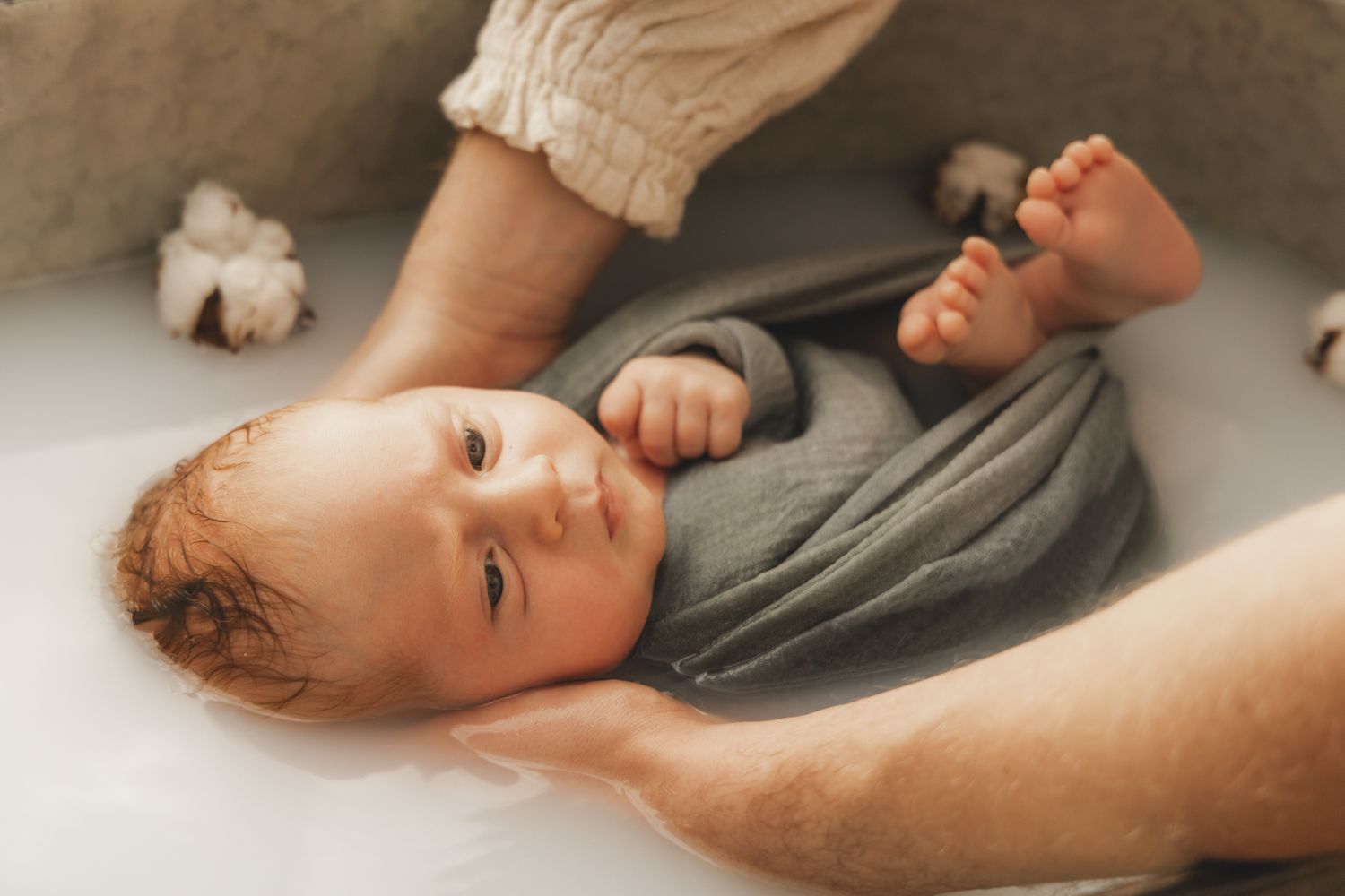 Newborn baby wrapped in gray cloth, gazing upward during a soothing milk bath.