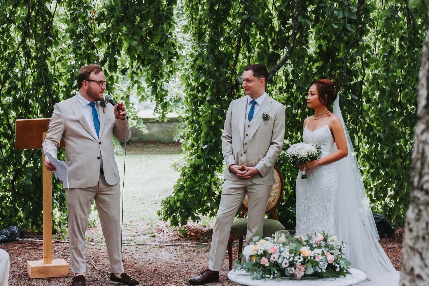 Outdoor wedding ceremony with officiant, groom in tan suit and bride under weeping willow tree.