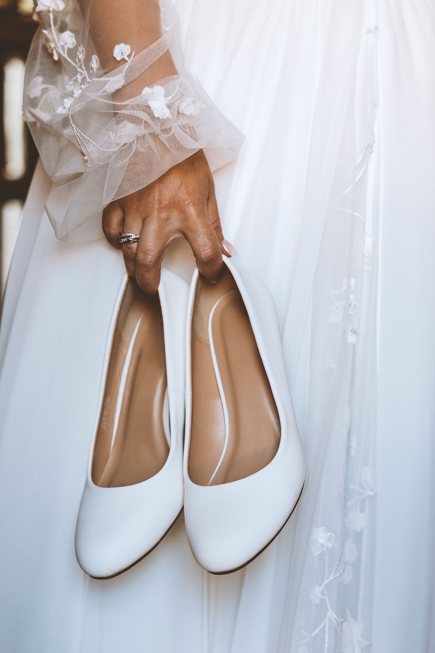 Bride holding elegant white heels with floral lace sleeve detail and diamond ring visible.