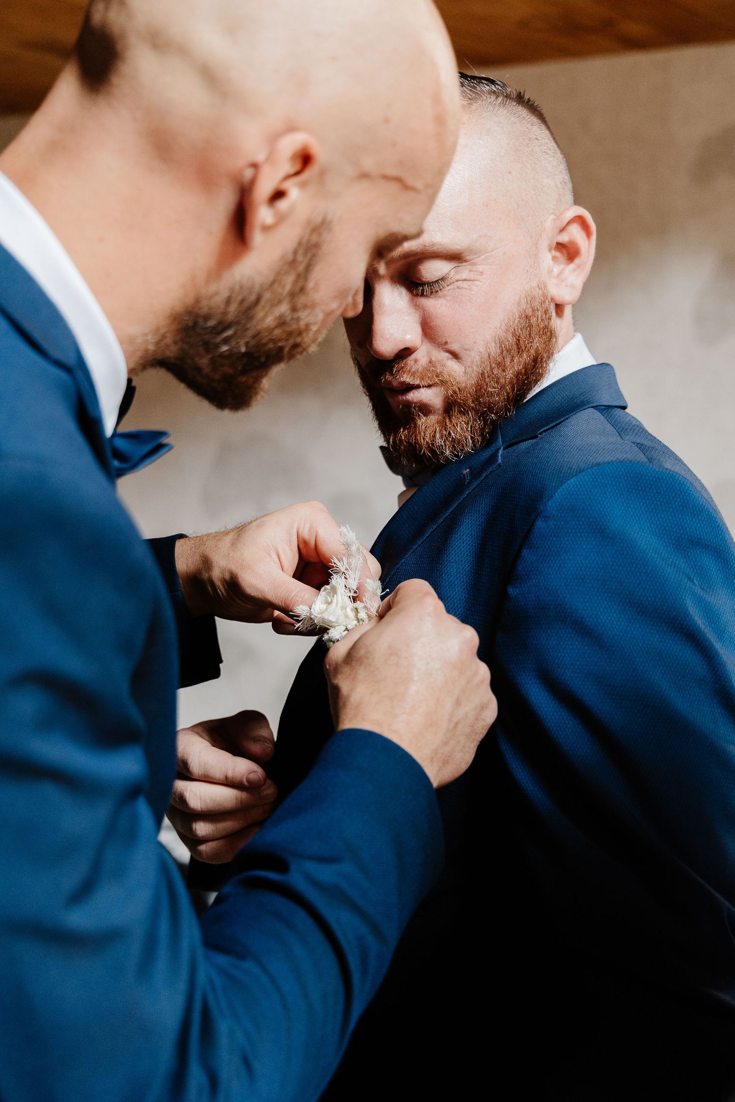 Two bearded men in navy suits, one pinning a white boutonniere on the other's lapel.