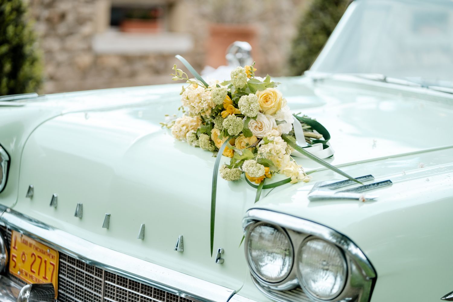 Wedding bouquet of white and yellow roses displayed on hood of vintage mint green classic car.
