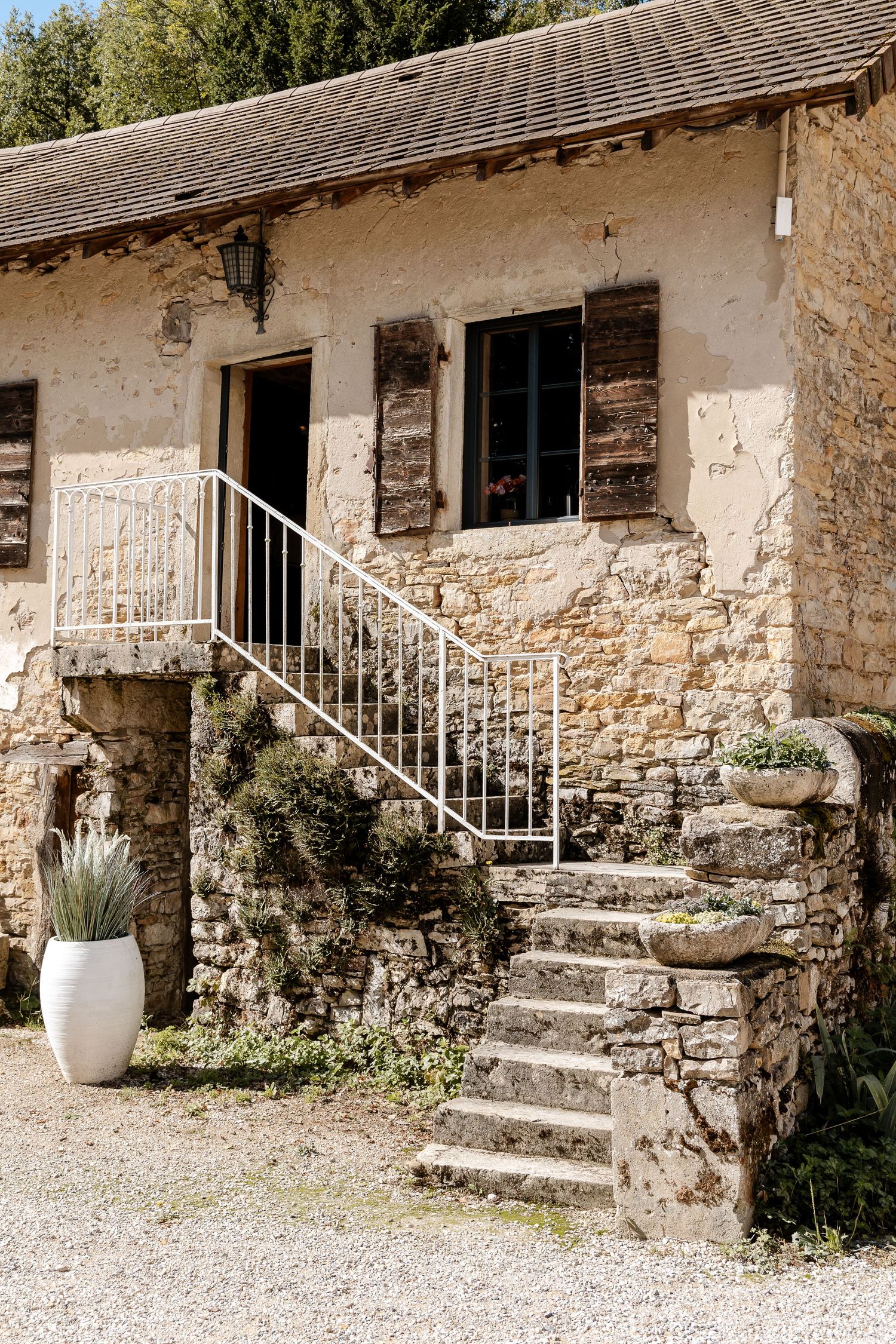 Rustic French stone farmhouse with wooden shutters, white iron staircase railing, and potted plants on gravel courtyard.