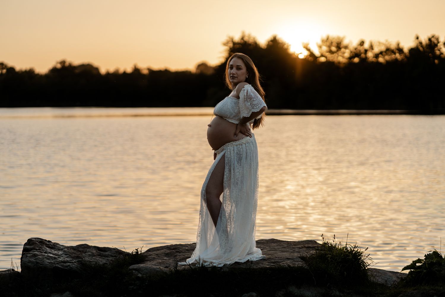 Pregnant woman in white dress standing on rocks by lake at golden sunset.