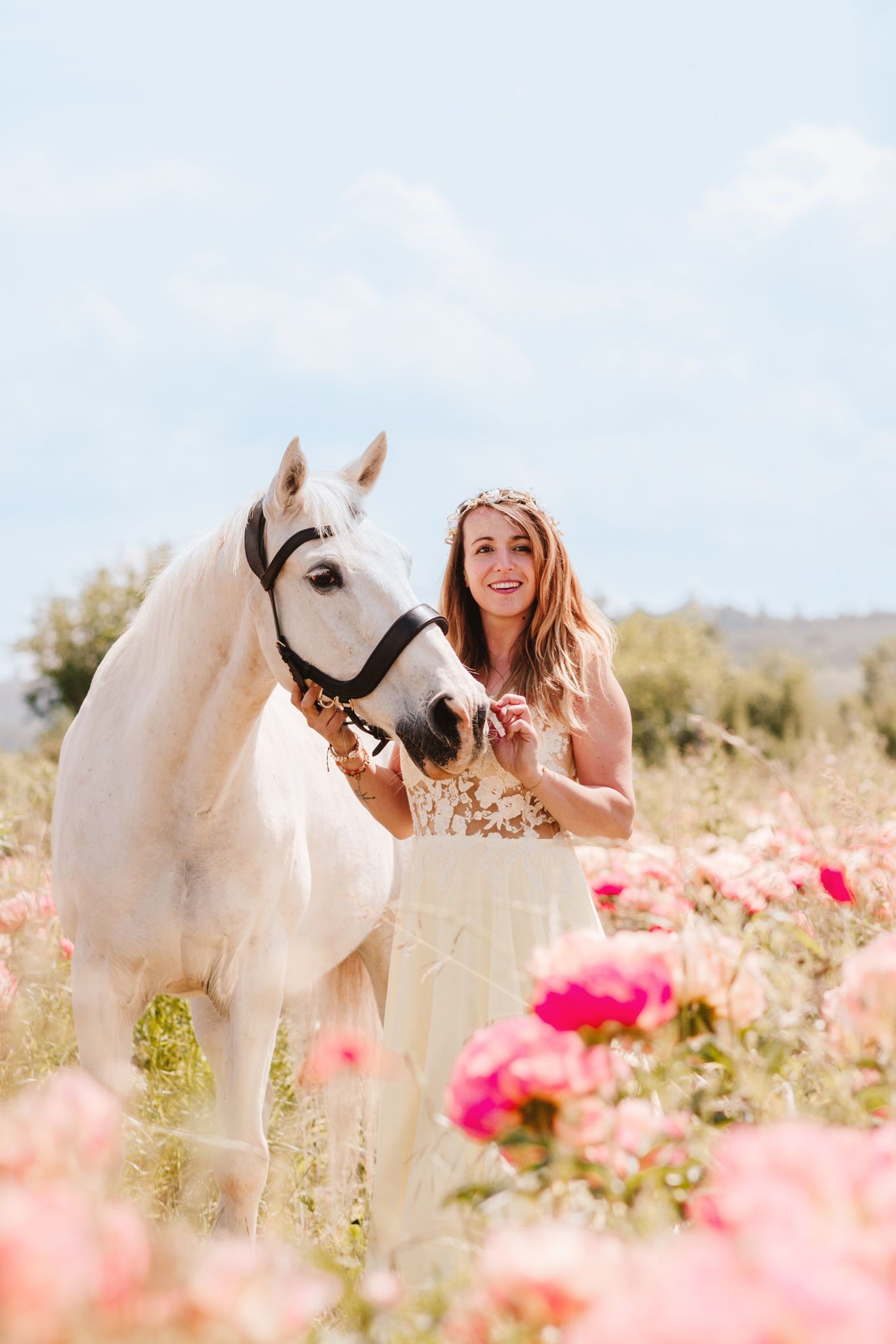 Happy woman in white dress standing beside white horse in pink peony field.
