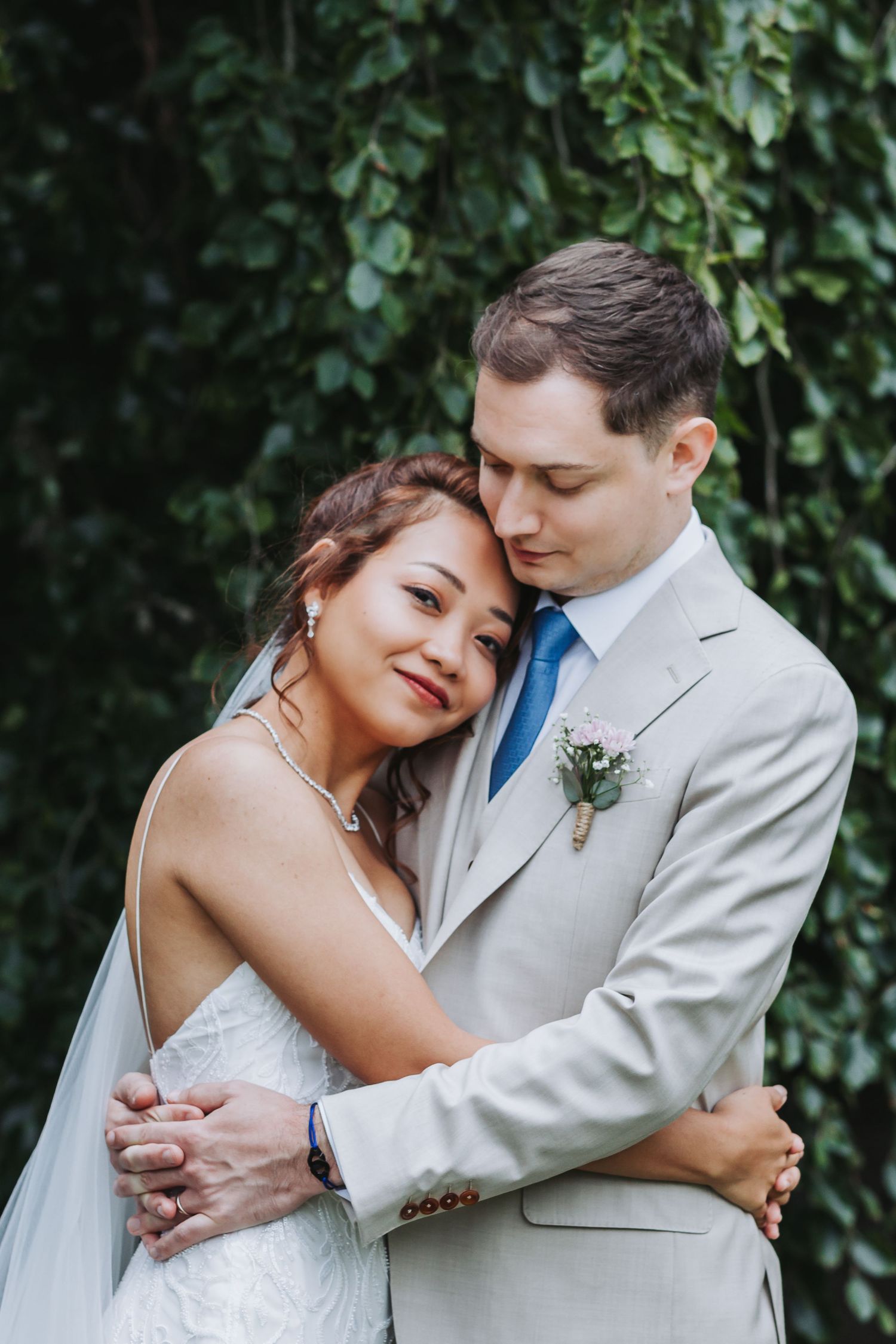 Bride and groom sharing tender embrace against lush green ivy wall, groom in beige suit with blue tie.