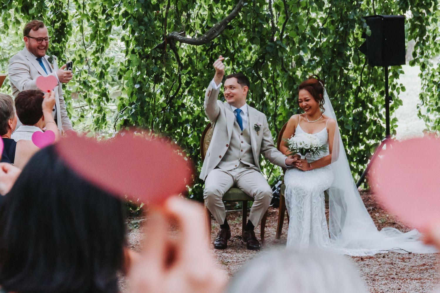 Joyful groom raising glass while seated with bride during outdoor wedding ceremony under green trees.