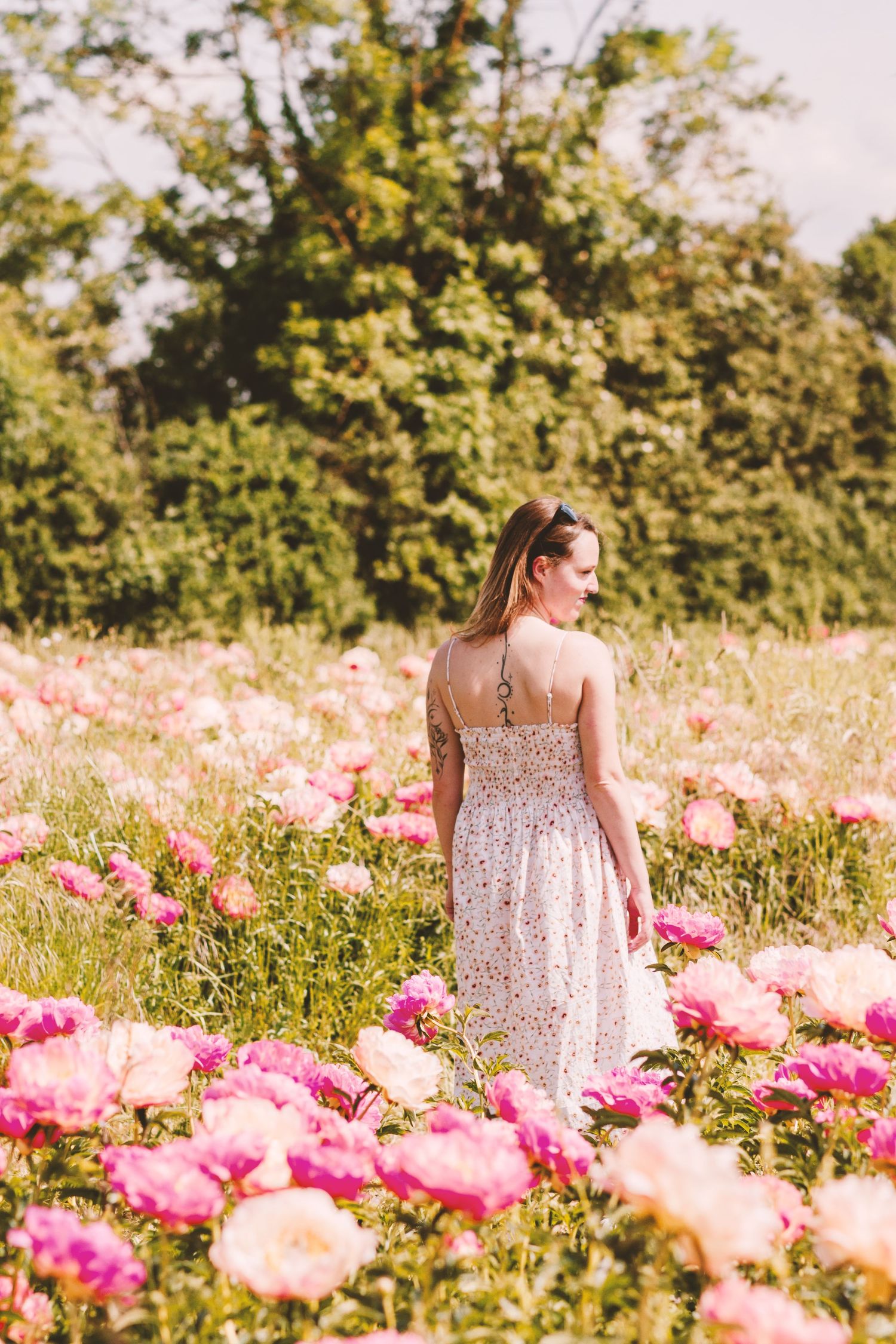 Young woman in white floral dress walking through a lush pink and magenta peony flower field.