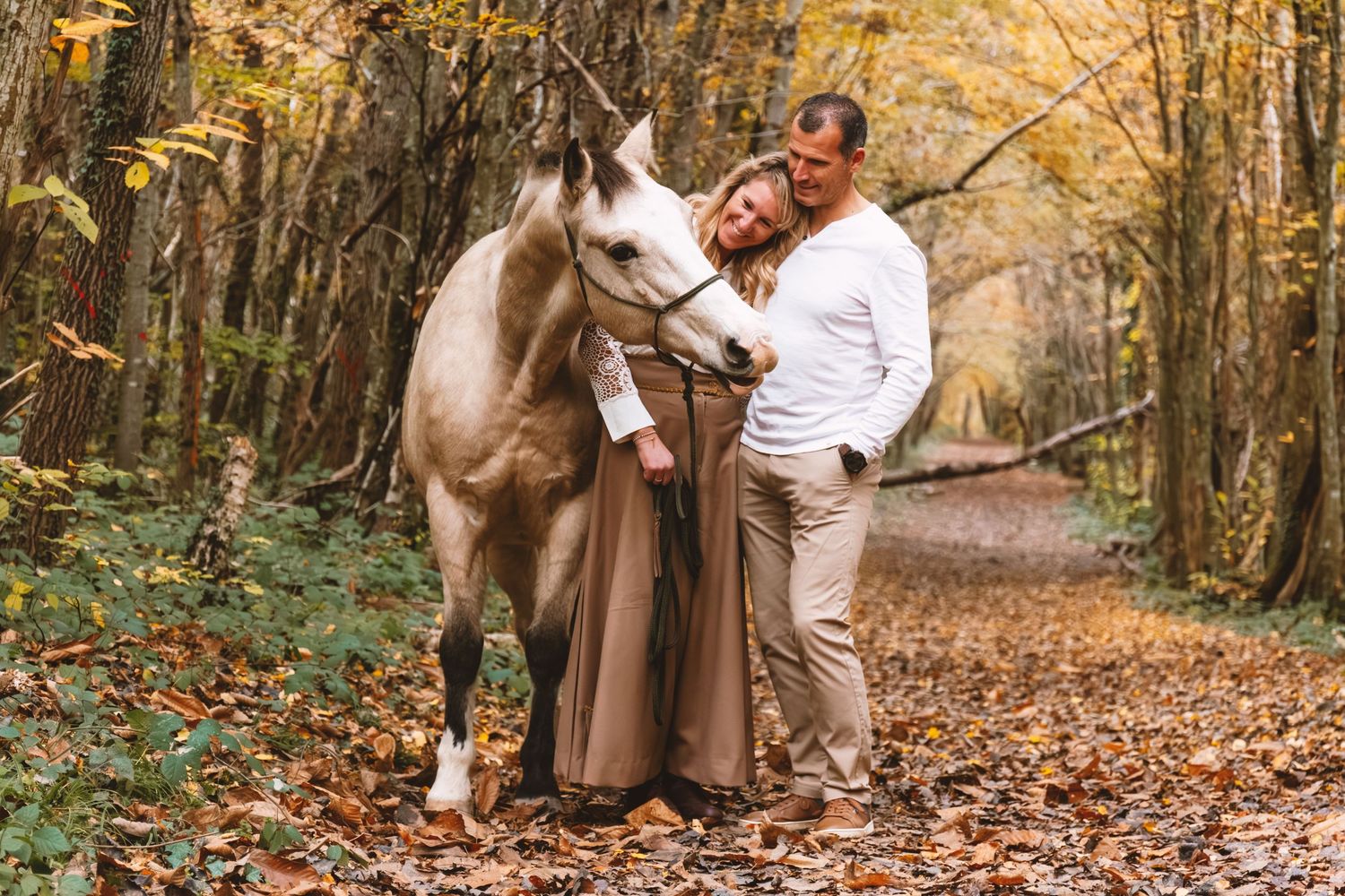 Woman in brown dress embracing white horse while man smiles on a leaf-covered forest trail.