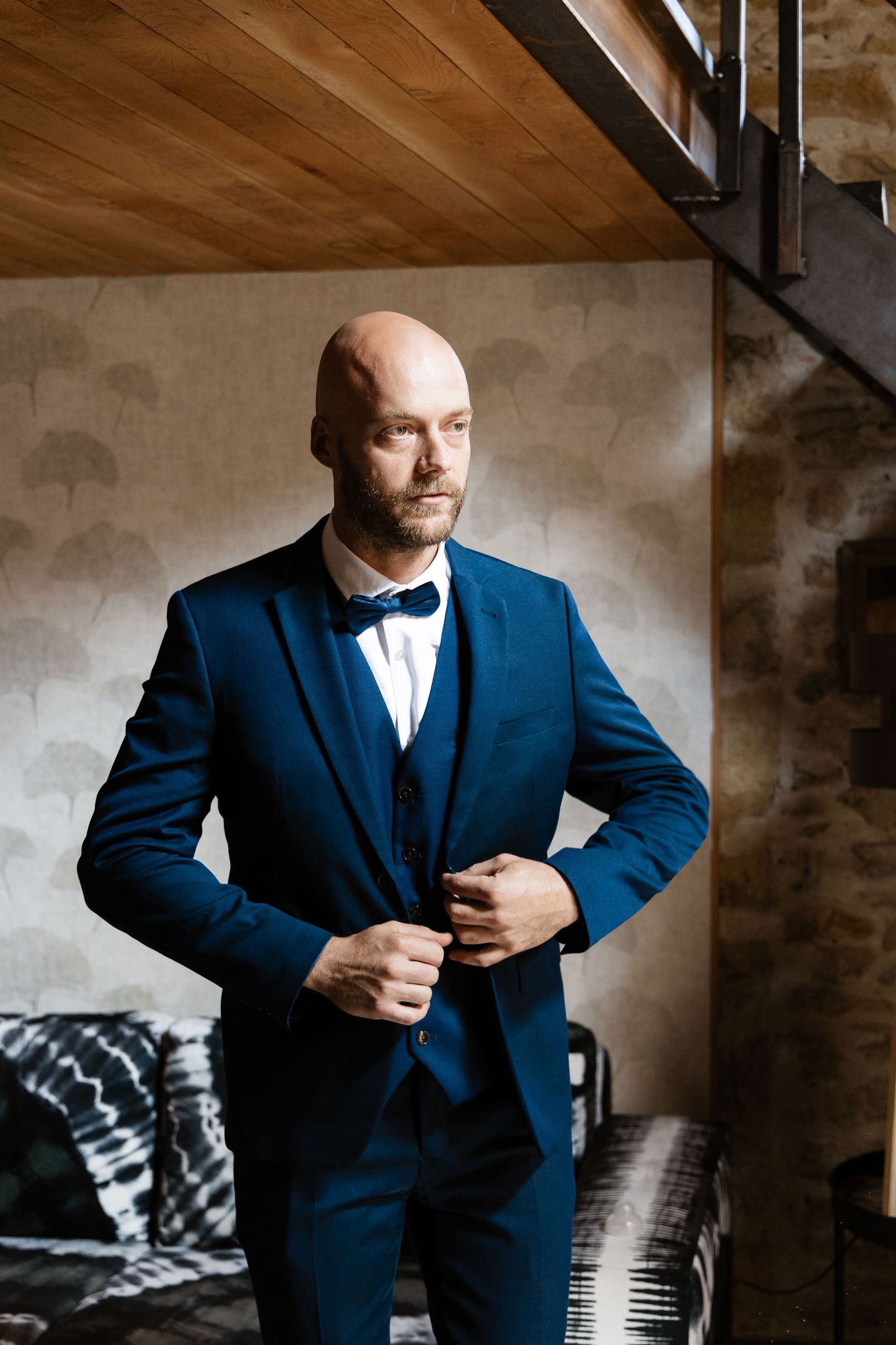 Bald groom in a navy blue three-piece suit and bow tie buttoning his jacket indoors.