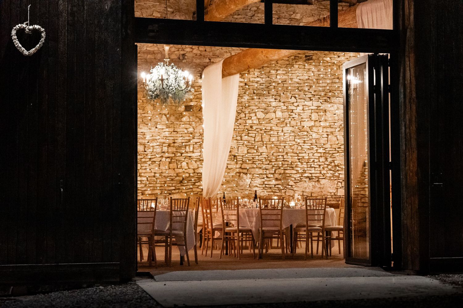 Rustic stone barn wedding venue entrance at night with chandelier and warm candlelit tables inside.