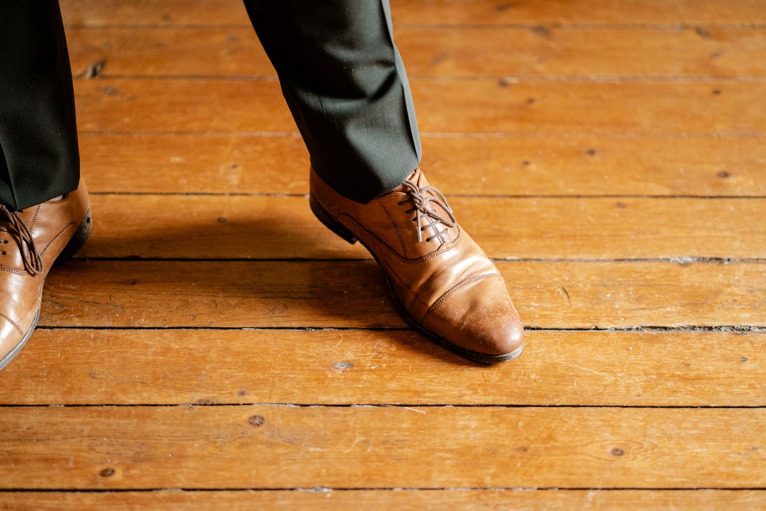 Close-up of groom's tan leather oxford shoes on rustic wooden floor.