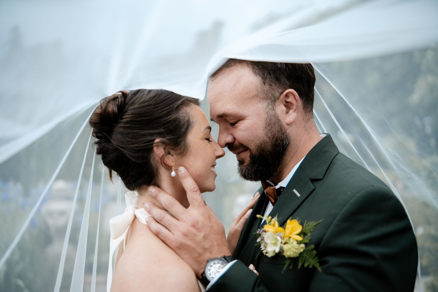 Bride and groom sharing intimate moment under wedding veil with yellow floral boutonniere.