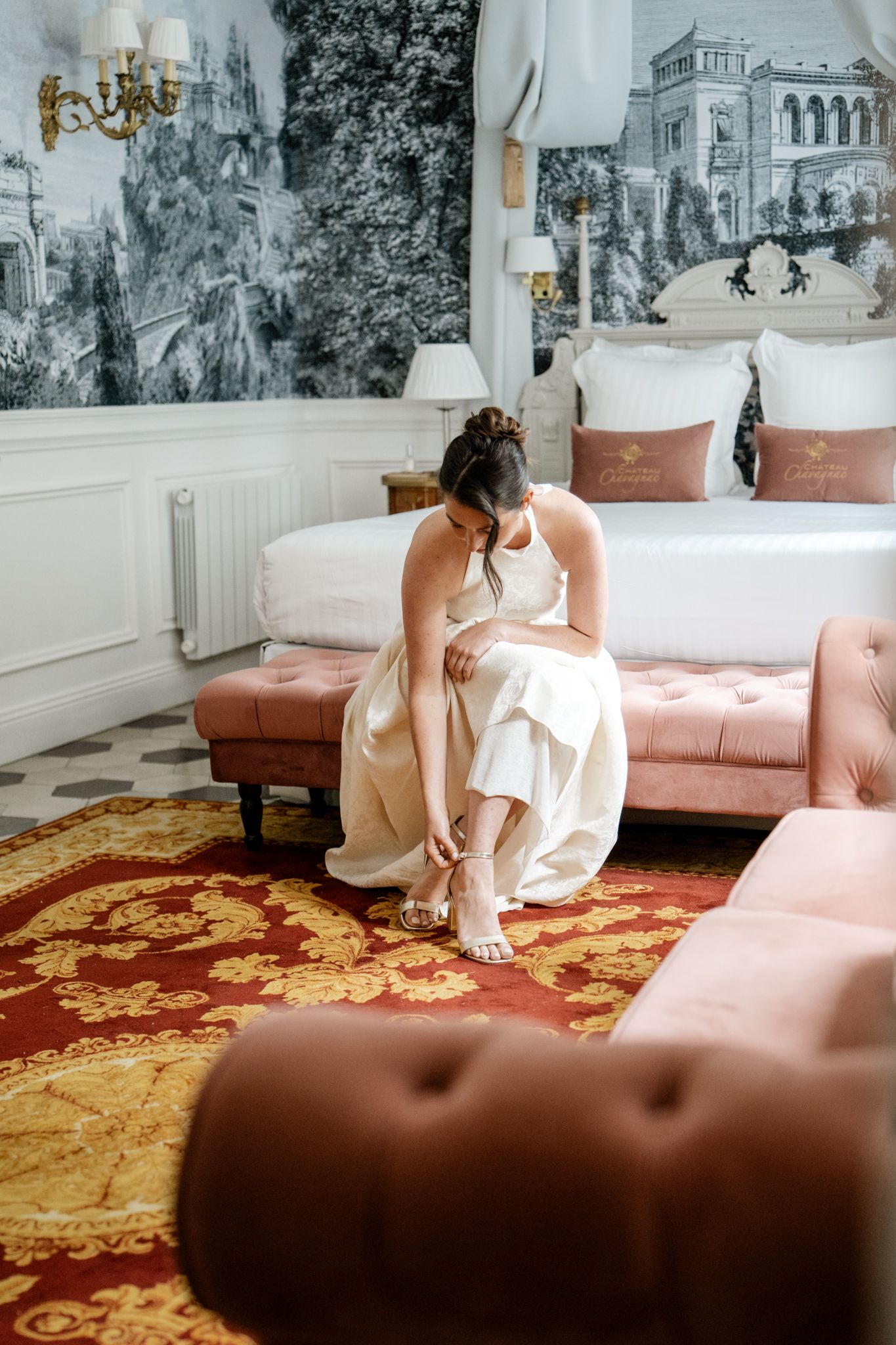 Bride in white gown sitting on pink velvet bench adjusting her wedding shoes in an elegant decorated hotel room.