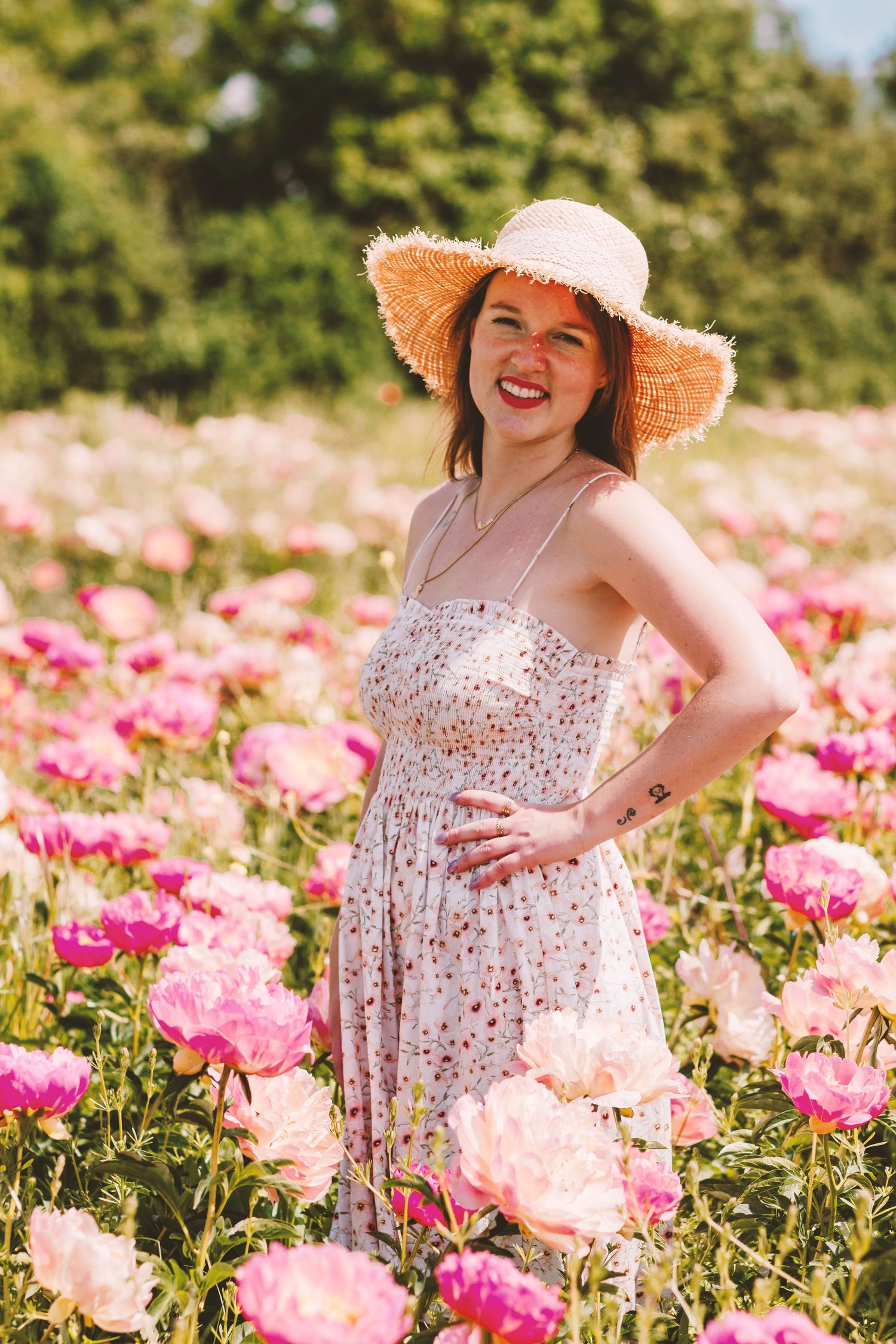 Smiling woman in floral dress and straw hat posing in pink peony field.