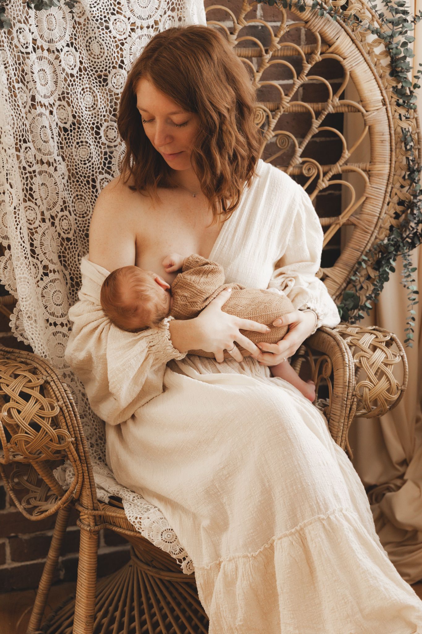 Mother in white dress breastfeeding newborn baby while sitting in a rattan peacock chair.