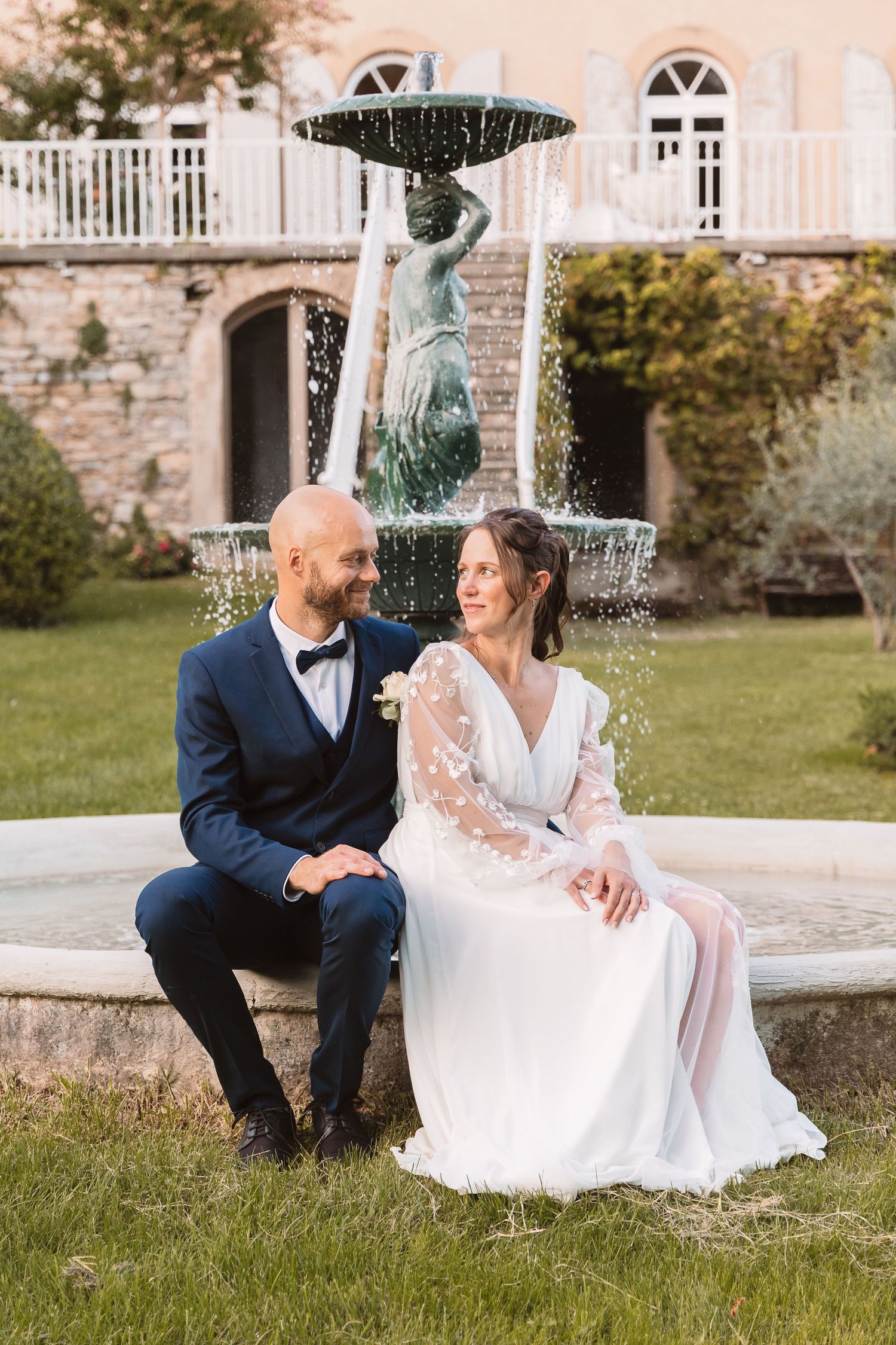 Bride and groom sitting by ornate stone fountain in elegant garden venue, bride in floral white gown.