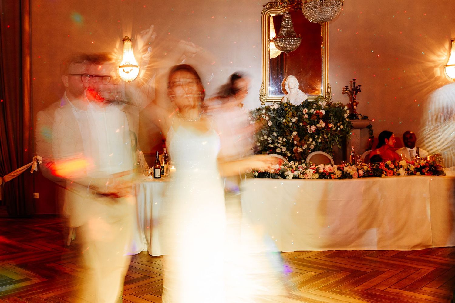 Bride dancing with guests in motion-blurred wedding reception with warm golden lighting.
