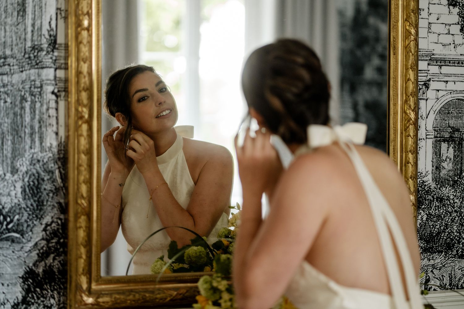 Bride in white halter gown smiling at her reflection in an ornate gold mirror while putting on earrings.