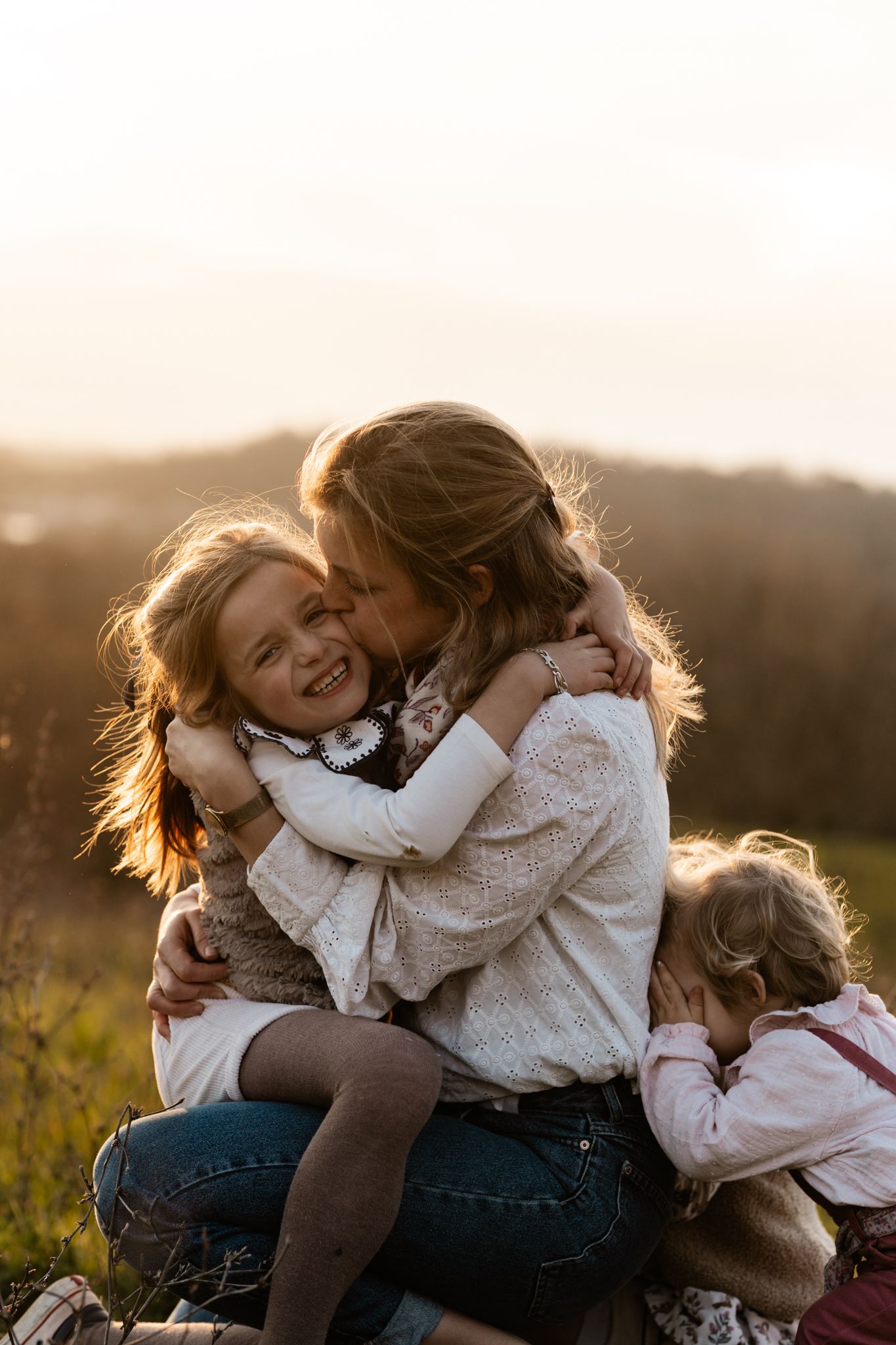 Séance photo famille en Isère : une mère enlace ses deux petites filles, dont l'une rit aux éclats, au coucher du soleil.