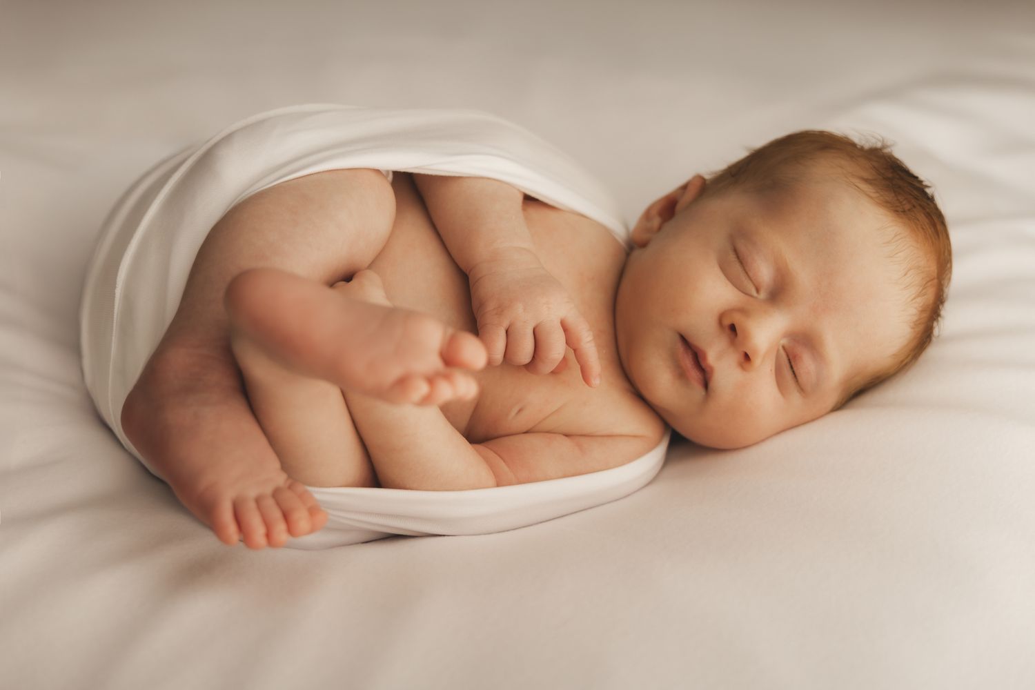 Newborn baby wrapped in white swaddle, sleeping peacefully on soft white surface.