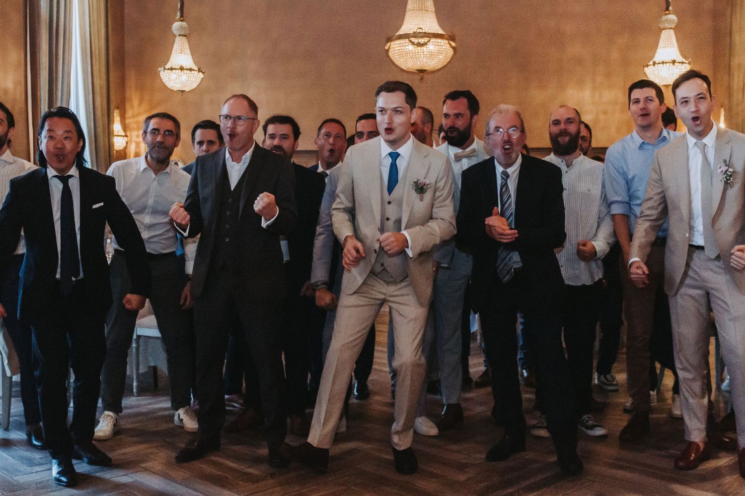 Excited groomsmen and male wedding guests dancing energetically in elegant chandelier ballroom.